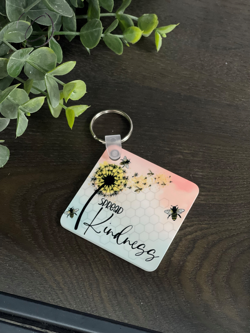 A vibrant double-sided keychain featuring the message 'Spread Kindness', made from durable plastic, measuring 2.25 inches square.