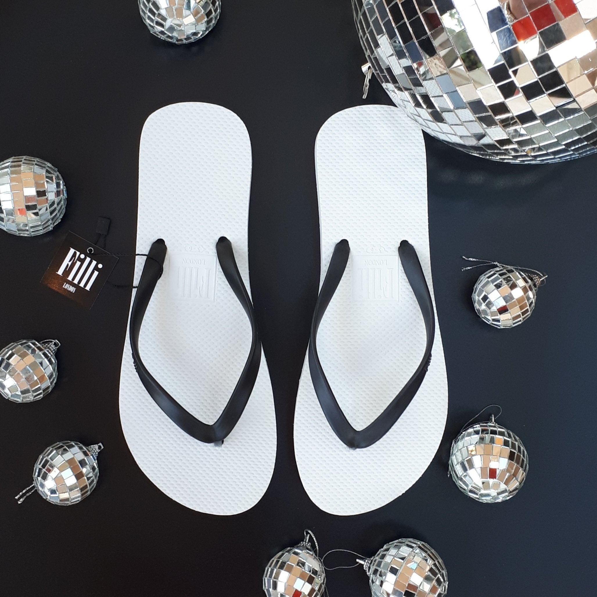 Filli London Naked Flip Flops in Black on White with embossed logo and rice pattern footbed, displayed in a cotton drawstring bag.