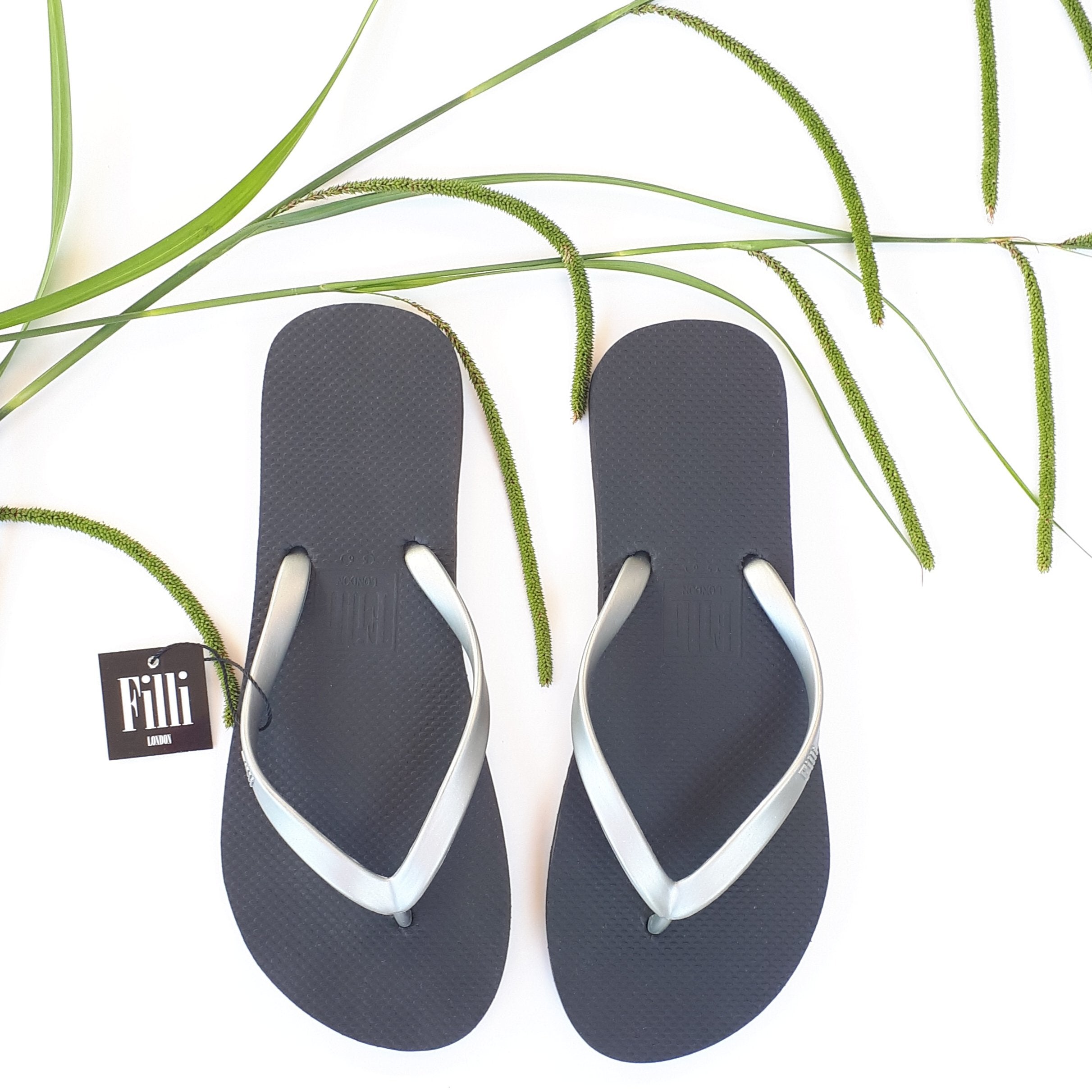 Filli London Naked Flip Flops in Silver on Black with embossed logo and rice pattern footbed, displayed in a cotton drawstring bag.