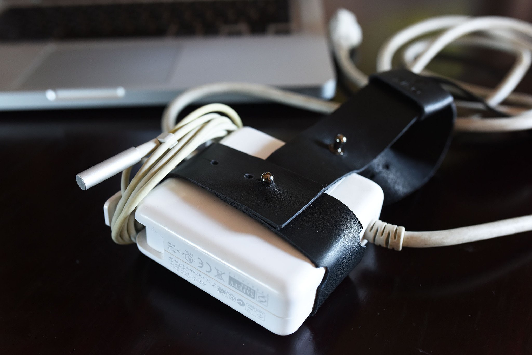 The Mac Cord Lord, a stylish leather cord organizer for MacBook chargers, featuring an adjustable cross-strap design.