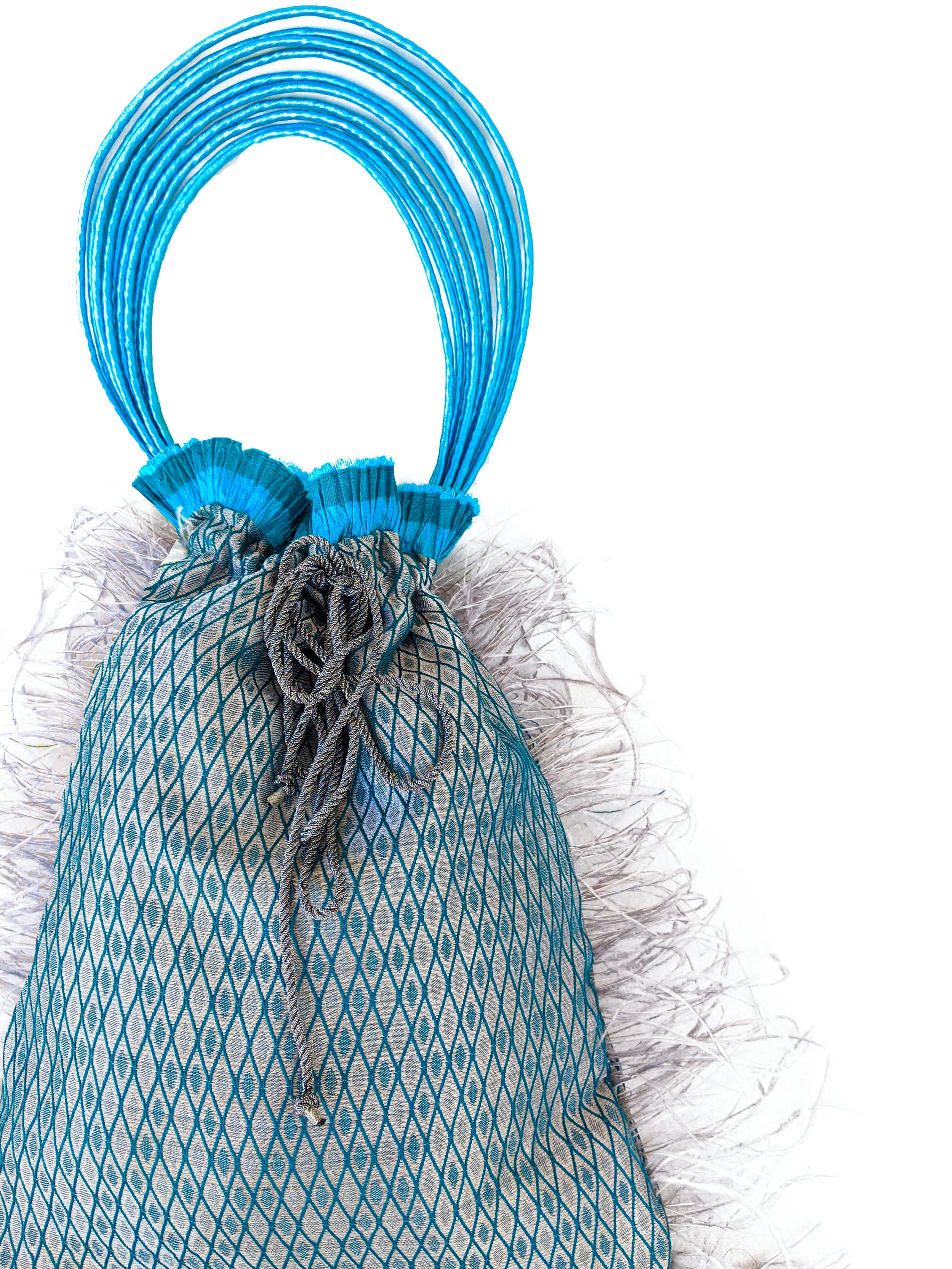 Imperial Jasper pouch bag featuring silvery blue fabric, turquoise accordion detail, grey feathers, and raffia cord handle.