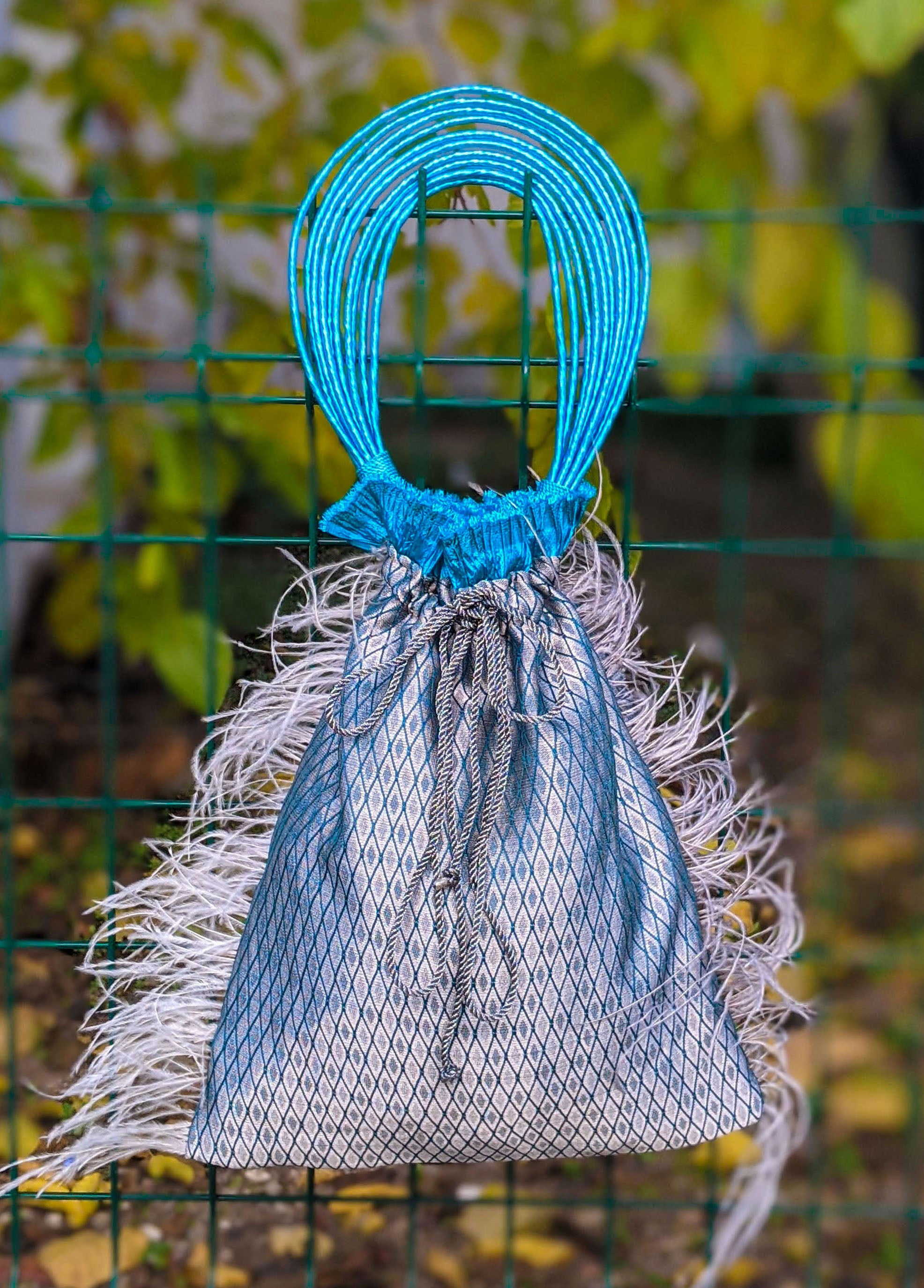 Imperial Jasper pouch bag featuring silvery blue fabric, turquoise accordion detail, grey feathers, and raffia cord handle.