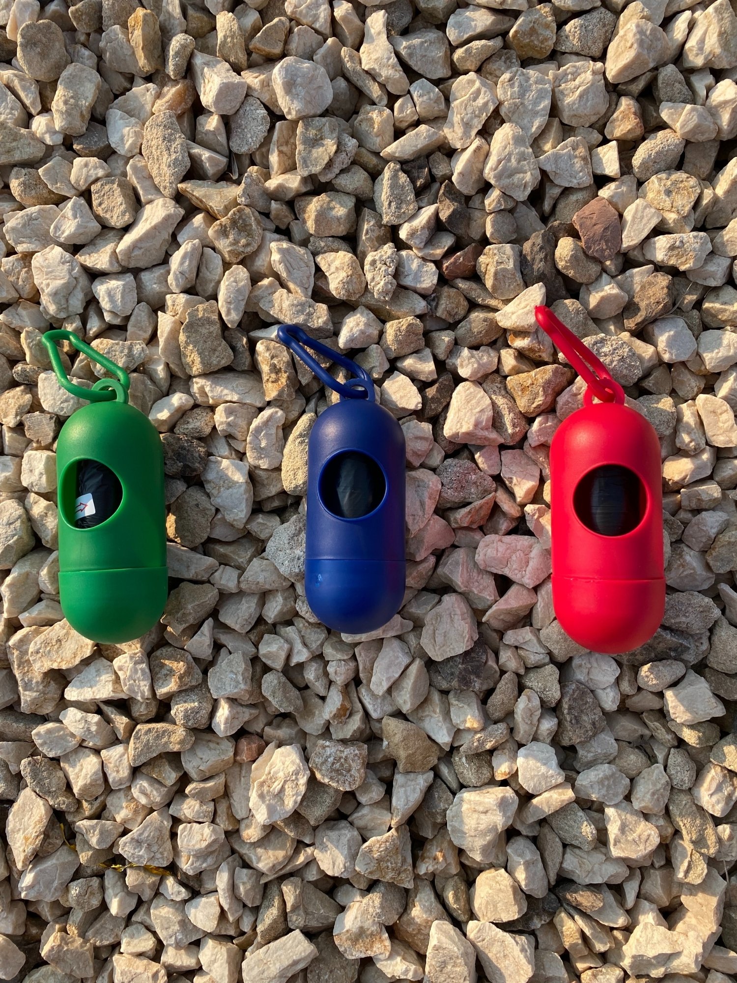 Dog Poop Bag Dispenser in Blue, Red, and Green colors with bags attached, showcasing its carabiner clip and easy-access slot.