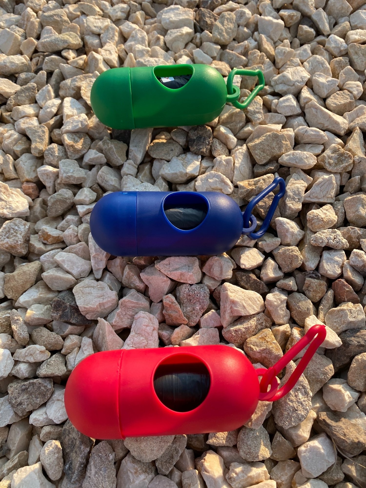 Dog Poop Bag Dispenser in Blue, Red, and Green colors with bags attached, showcasing its carabiner clip and easy-access slot.