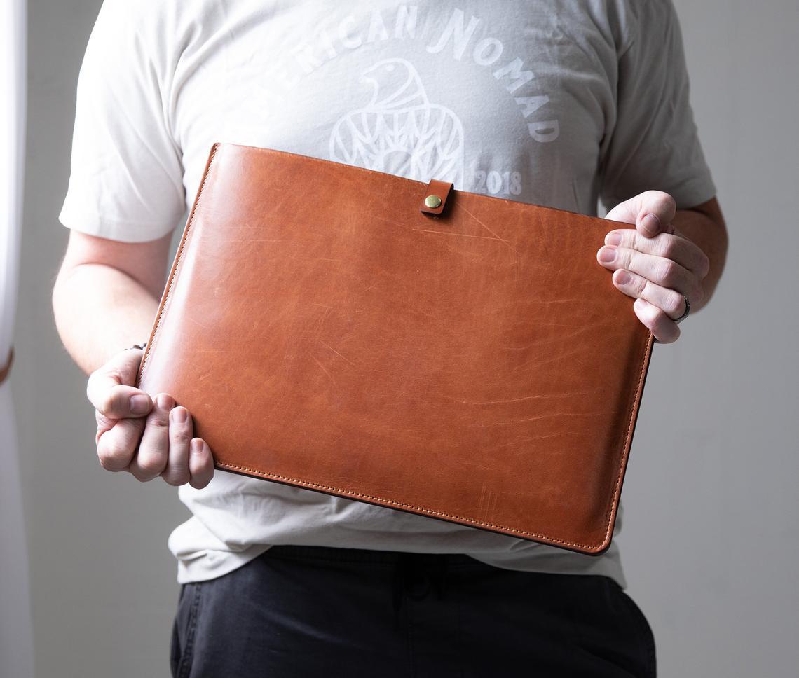 Personalized leather laptop sleeve made from genuine full grain leather, featuring a secure strap and brass snap, ideal for 13"-15" laptops.