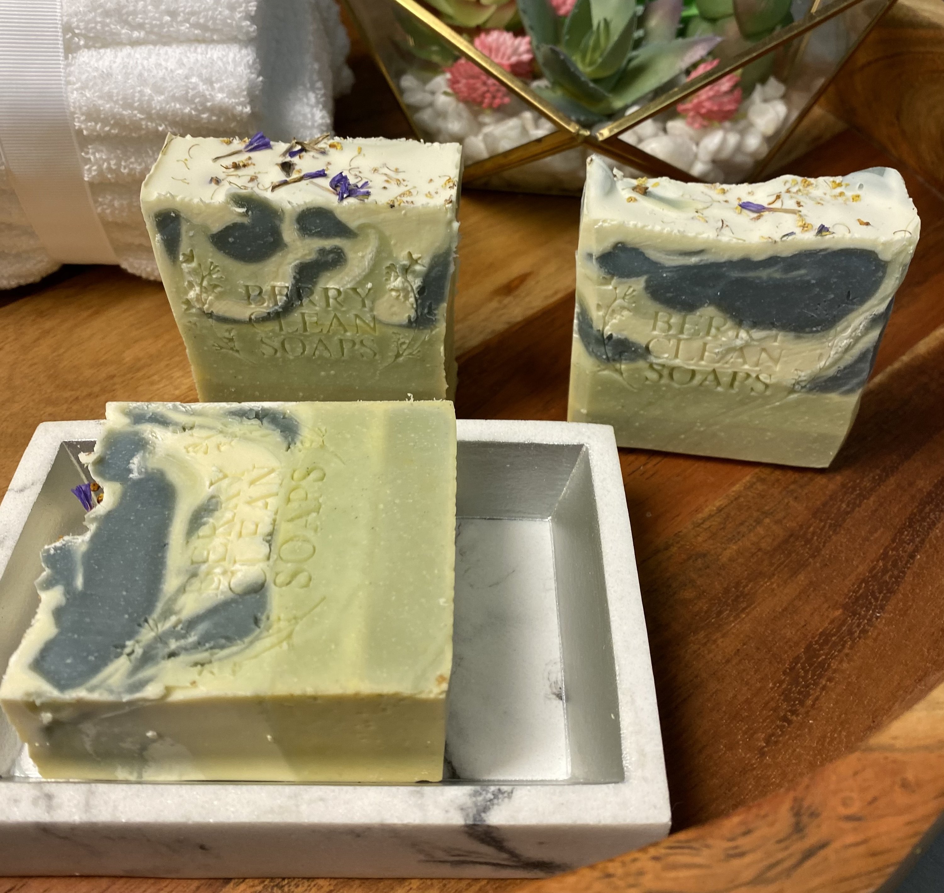 A beautifully crafted indigo soap bar showcasing a blend of blue and green hues, with visible spices and fruit elements, resting on a wooden surface.