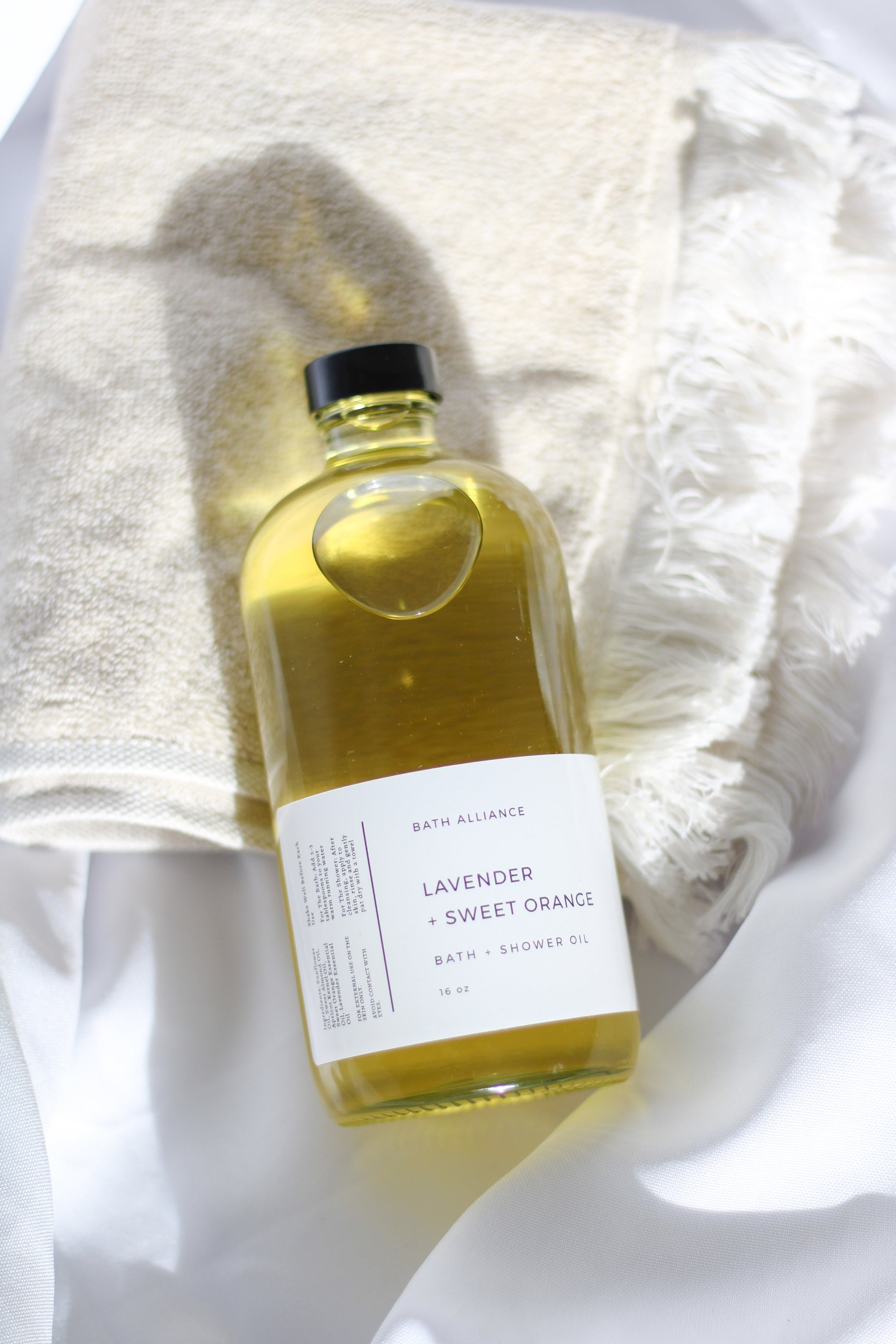 A 16 oz glass bottle of Lavender + Sweet Orange Bath + Shower Oil, showcasing its rich, golden color and elegant label design.