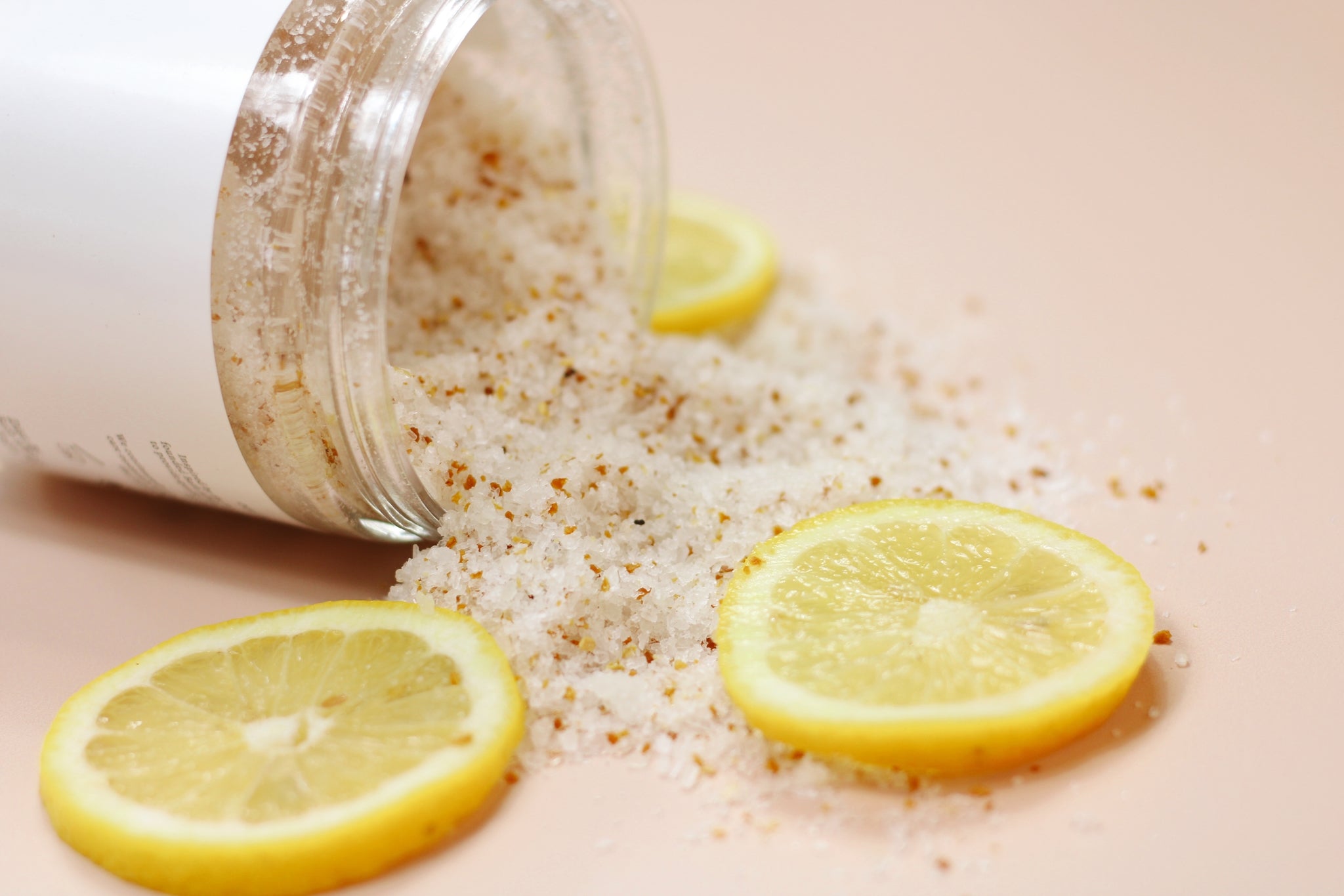 A jar of Lemon + Vanilla Bath Salt with a refreshing lemon and vanilla scent, surrounded by lemon slices and flowers.