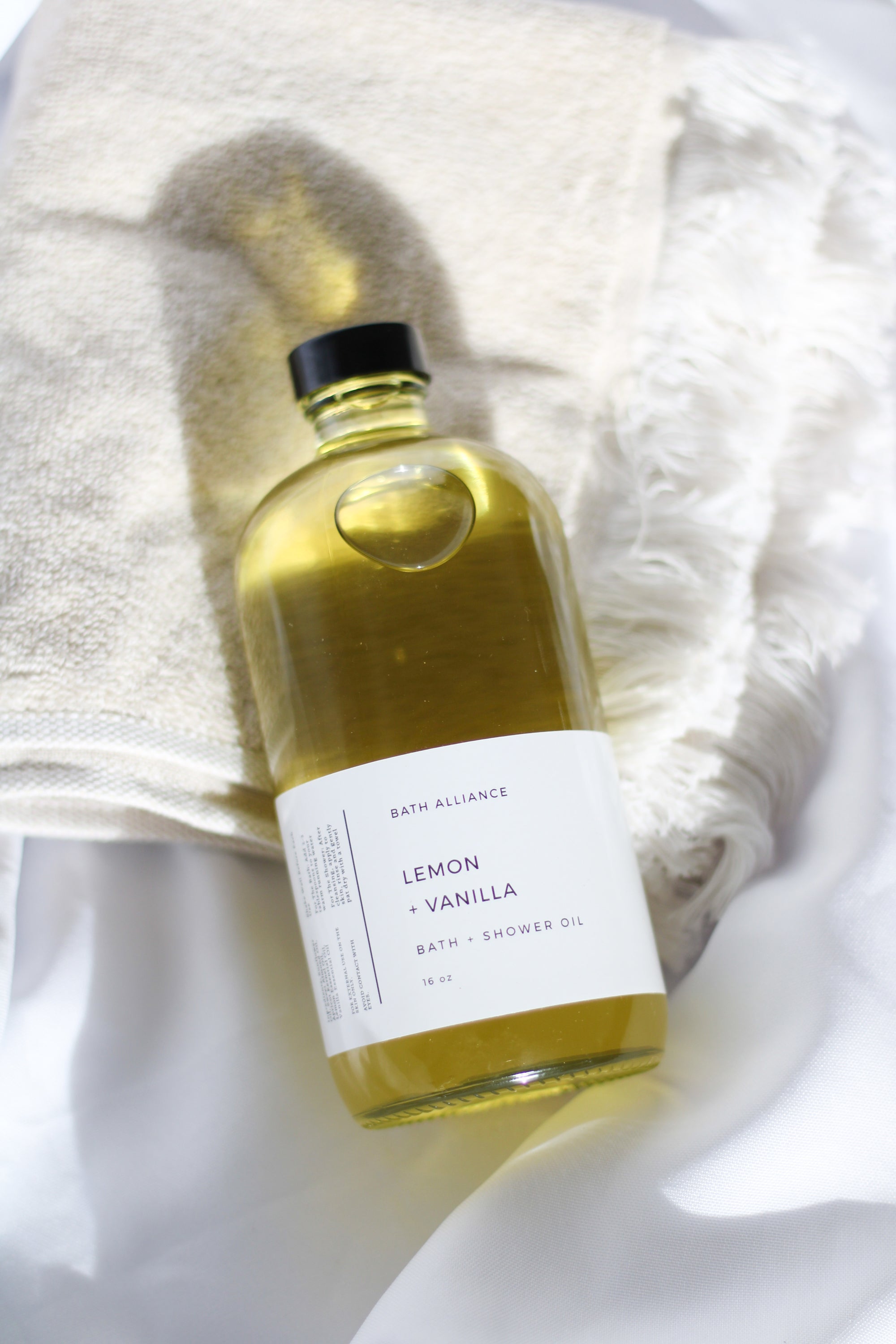 A 16 oz glass bottle of Lemon + Vanilla Bath + Shower Oil with a bright yellow label, showcasing its nourishing ingredients and refreshing aroma.