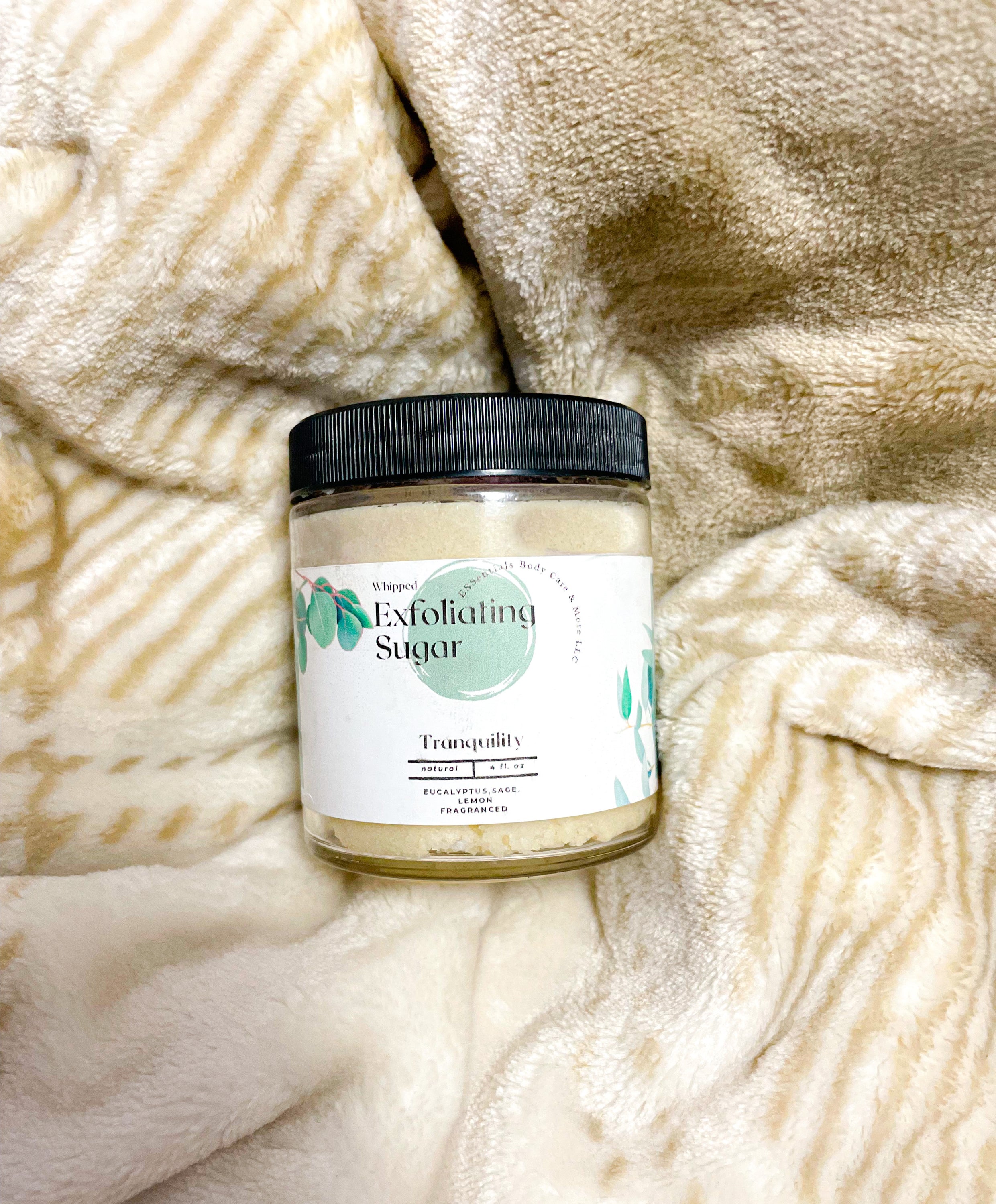 Tranquil exfoliating sugar with organic oils and butters in a clear jar, surrounded by natural ingredients like coconut and eucalyptus.