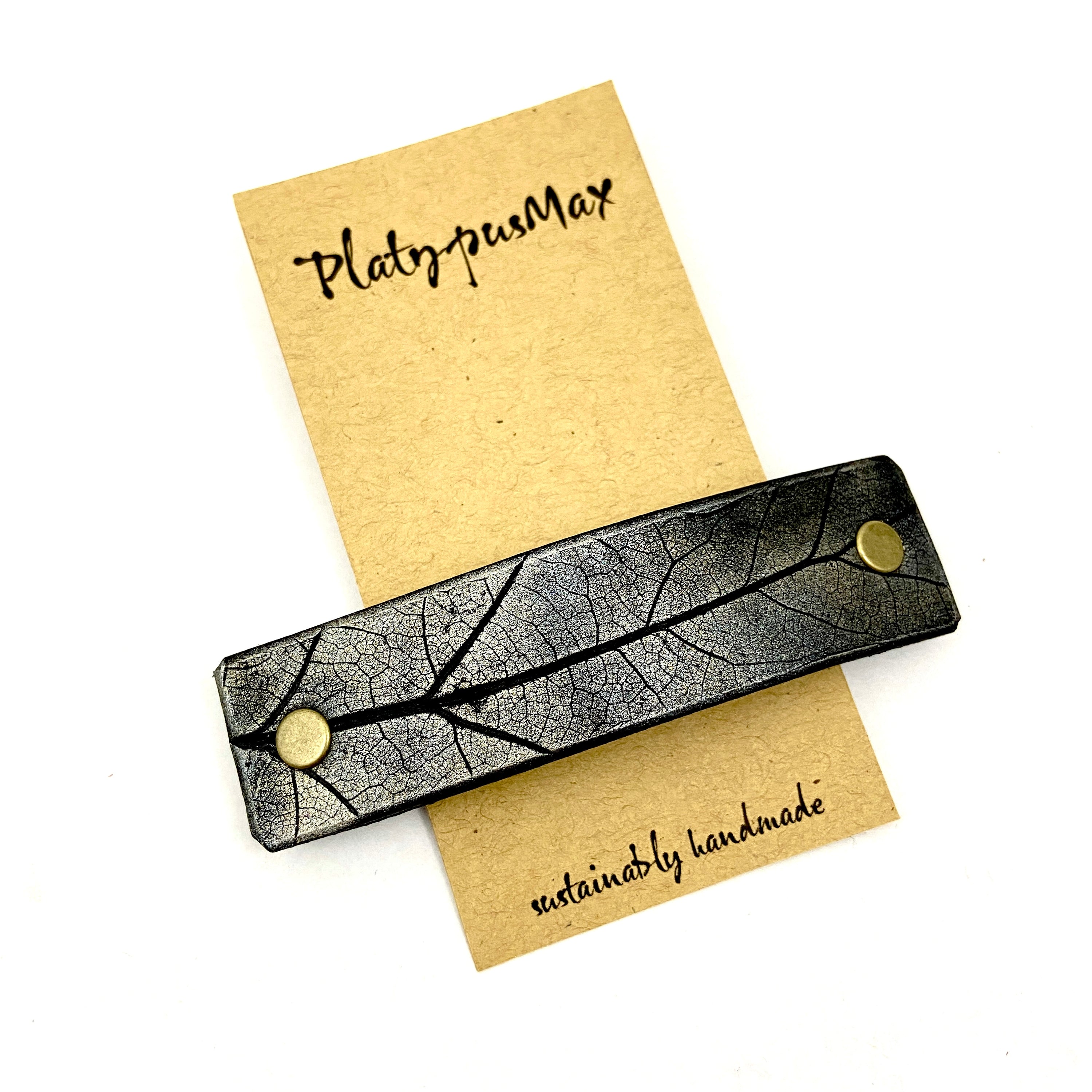 A handcrafted platinum oak leaf pressed leather hair barrette featuring an embossed leaf design on black-dyed leather.