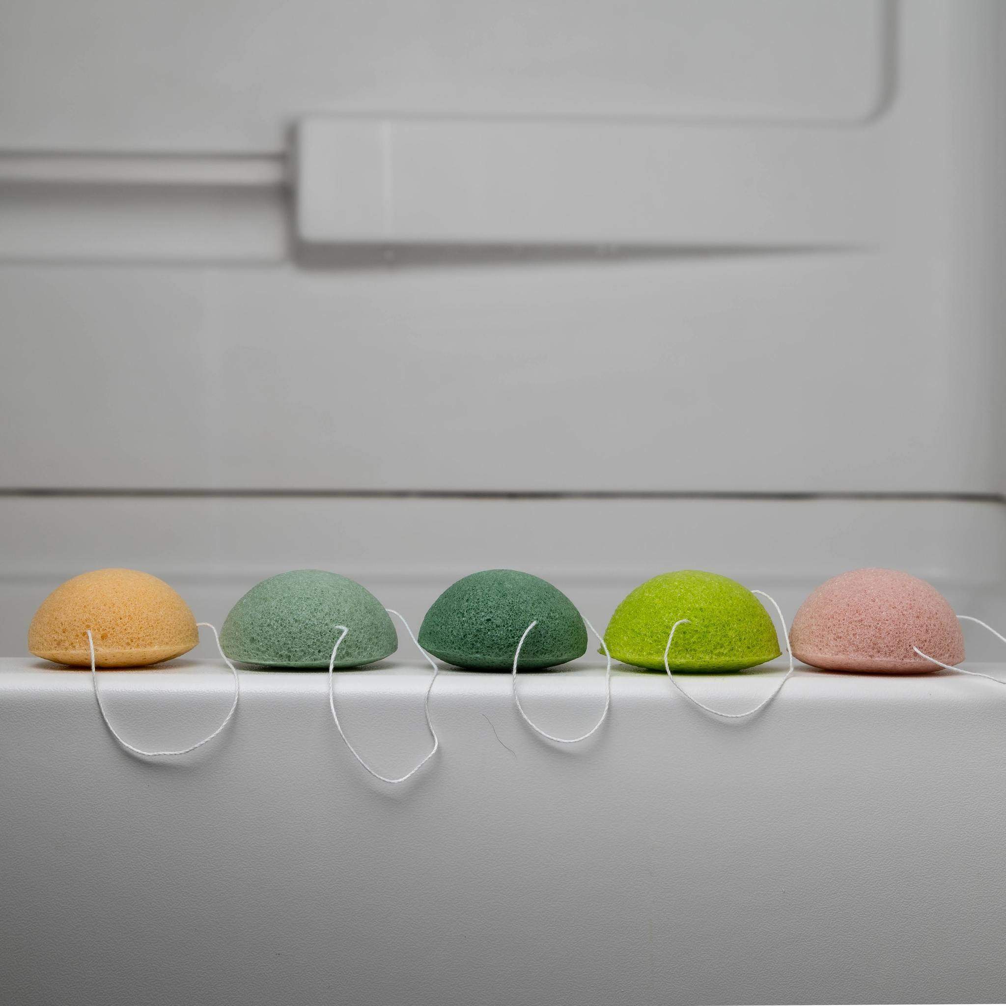 A collection of five biodegradable Konjac sponges in various colors, each representing different skin benefits, displayed on a natural wooden surface.