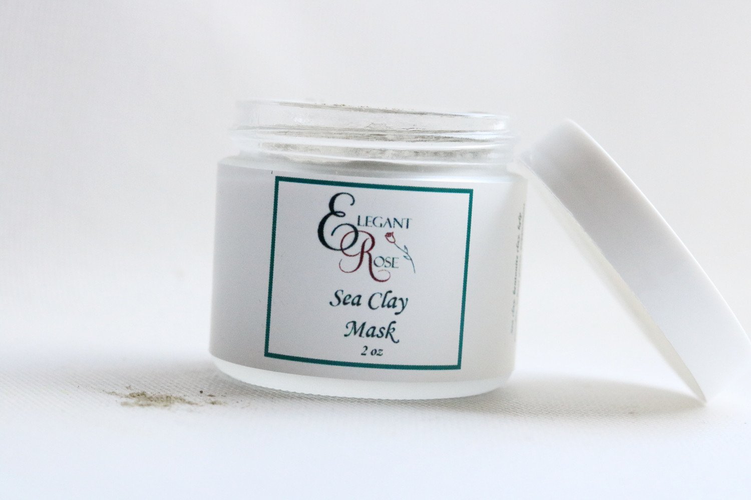 A 2-ounce glass jar of Sea Clay Facial Mask, showcasing its natural powder texture, surrounded by fresh ingredients like wheat grass and kelp powder.