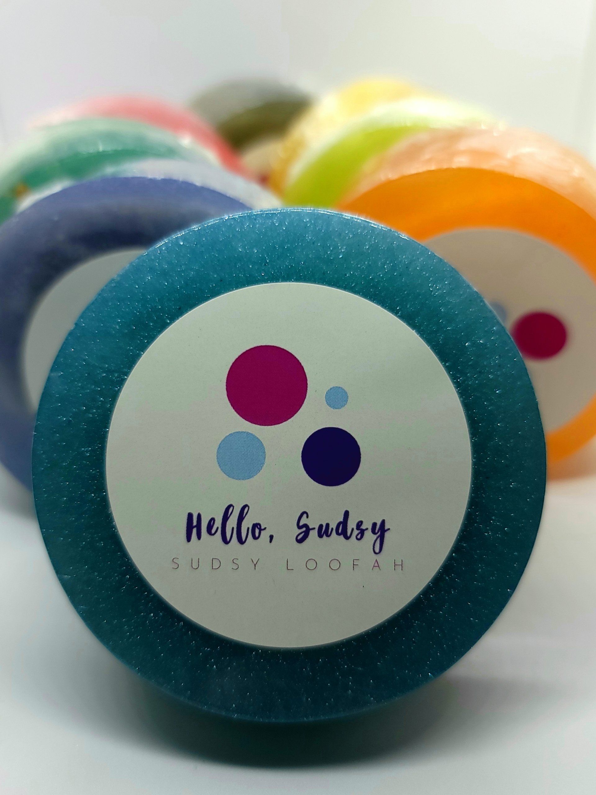 A collection of Sudsy Loofahs in various scents, showcasing their texture and vibrant colors, perfect for exfoliating and moisturizing skin.