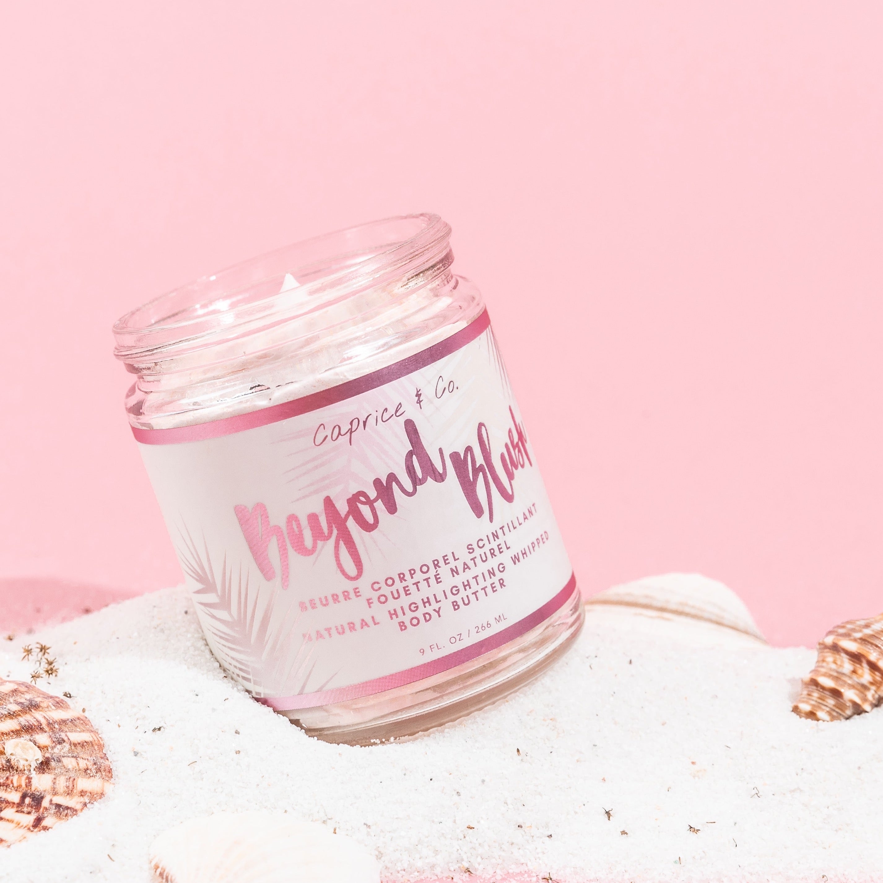 A jar of Beyond Blush Highlighting Body Butter with a rose gold hue, surrounded by tropical flowers and coconuts, showcasing its luxurious texture.