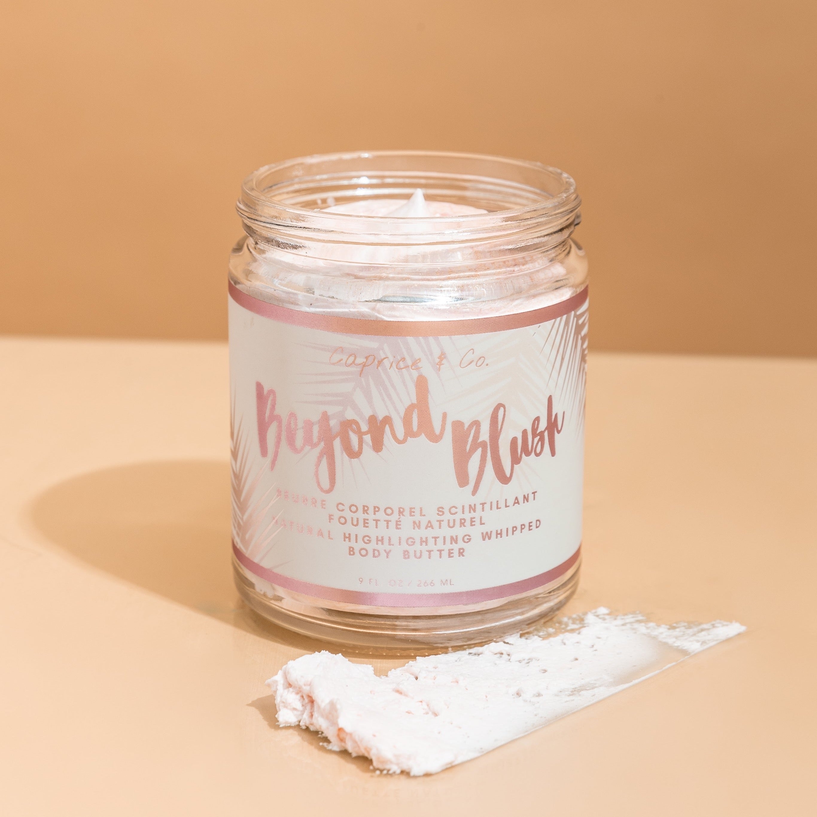 A jar of Beyond Blush Highlighting Body Butter with a rose gold hue, surrounded by tropical flowers and coconuts, showcasing its luxurious texture.