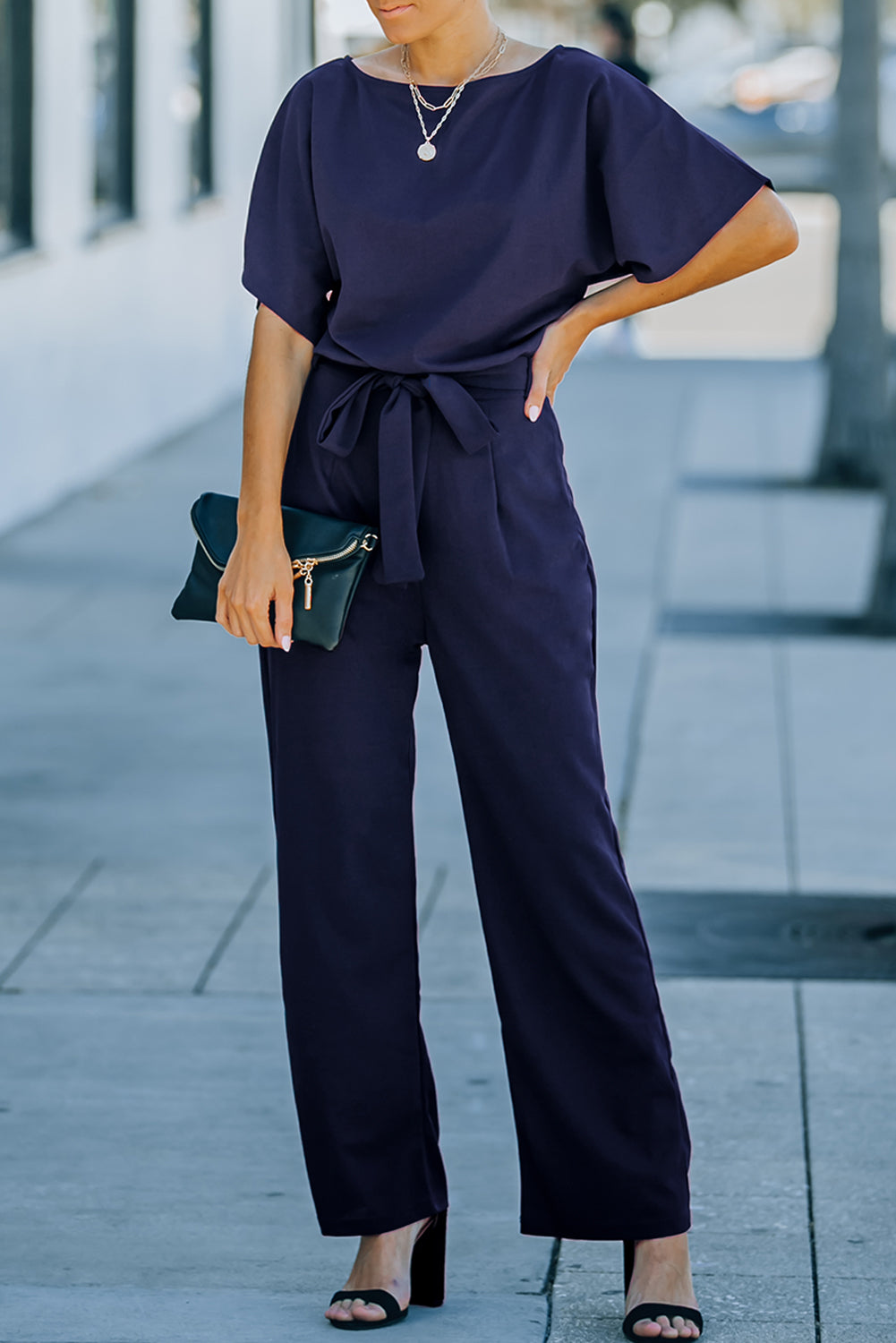 A stylish woman wearing a blue belted wide leg jumpsuit with batwing sleeves, showcasing a chic and glamorous look.