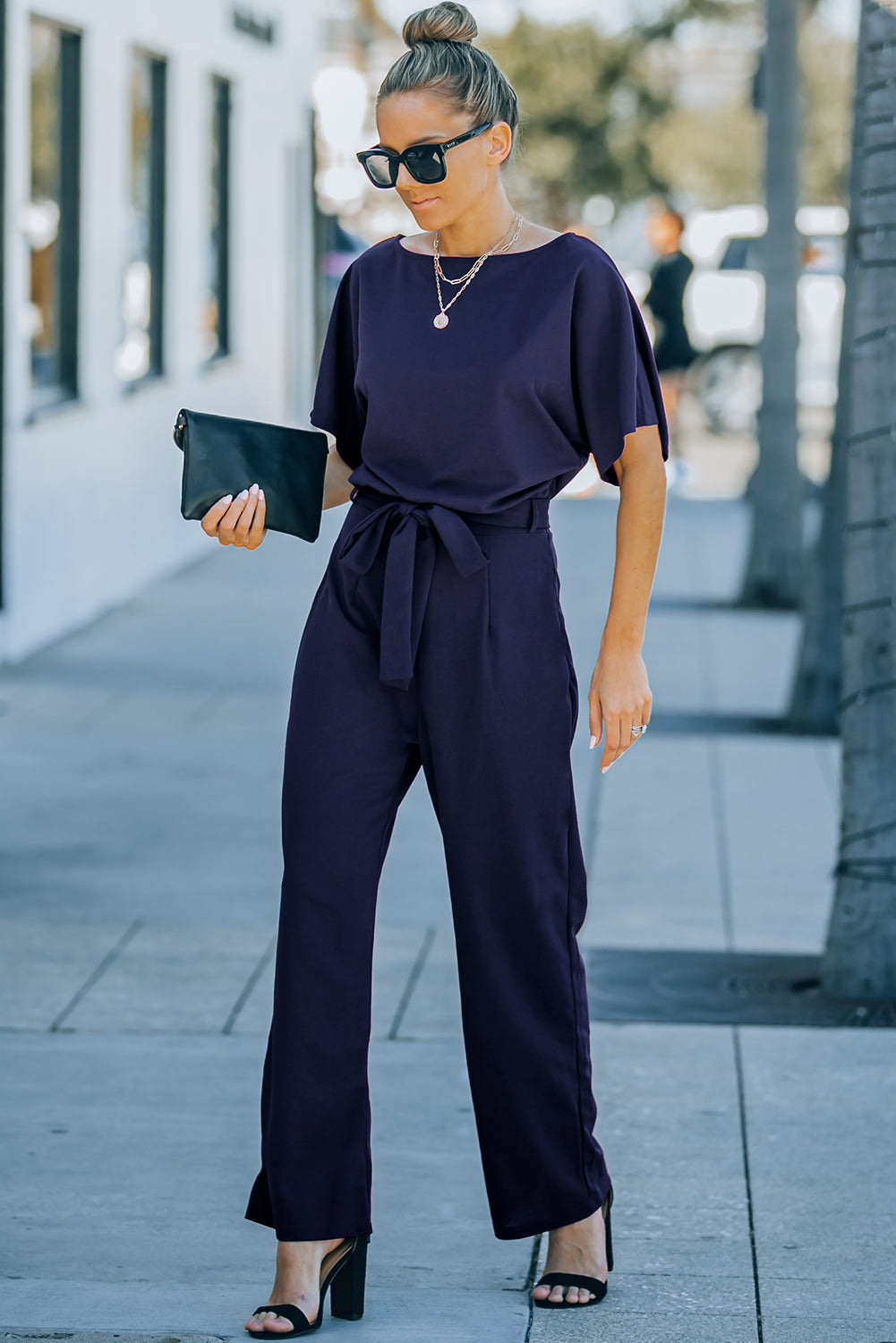 A stylish woman wearing a blue belted wide leg jumpsuit with batwing sleeves, showcasing a chic and glamorous look.