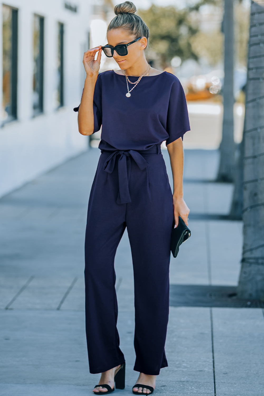 A stylish woman wearing a blue belted wide leg jumpsuit with batwing sleeves, showcasing a chic and glamorous look.