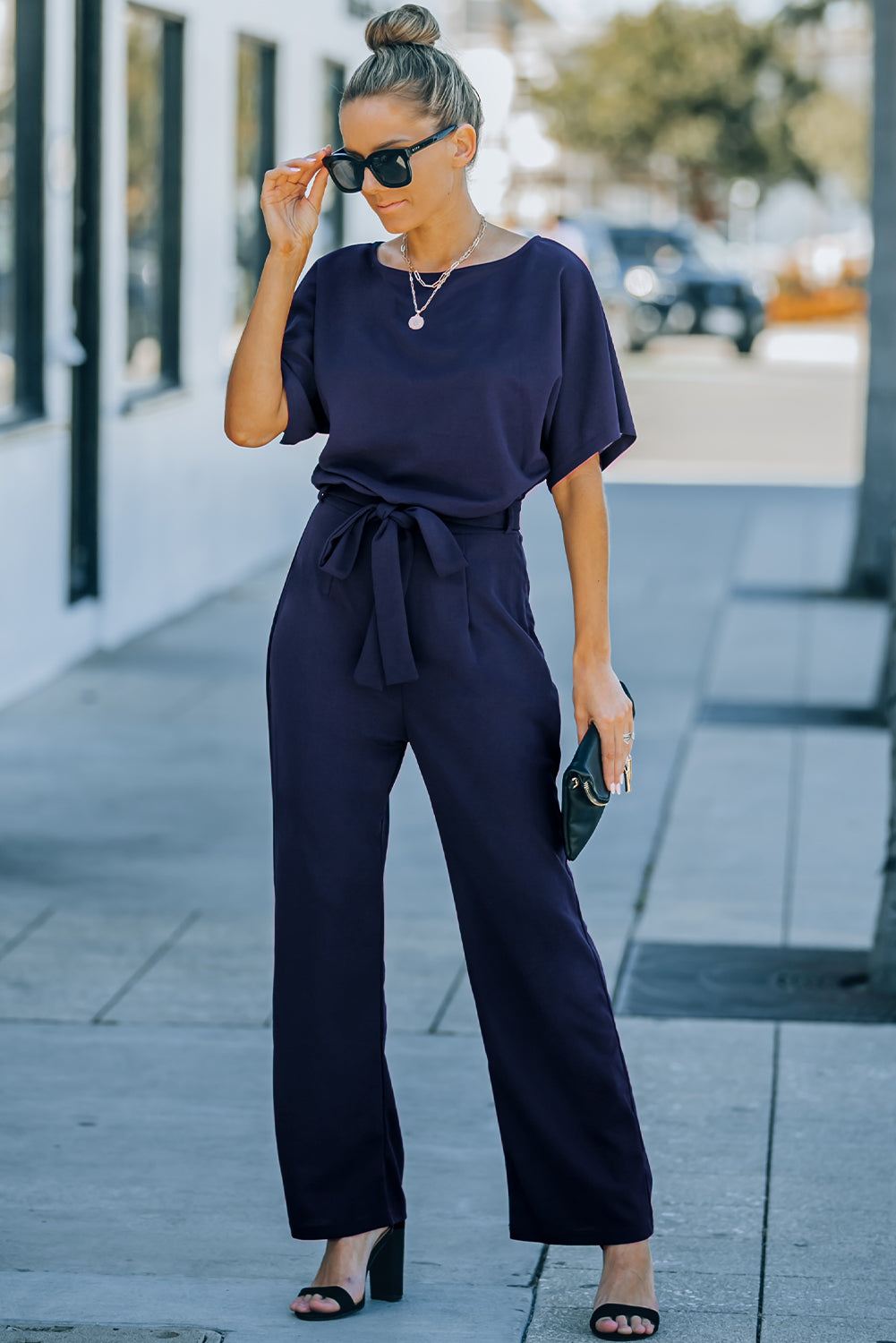 A stylish woman wearing a blue belted wide leg jumpsuit with batwing sleeves, showcasing a chic and glamorous look.