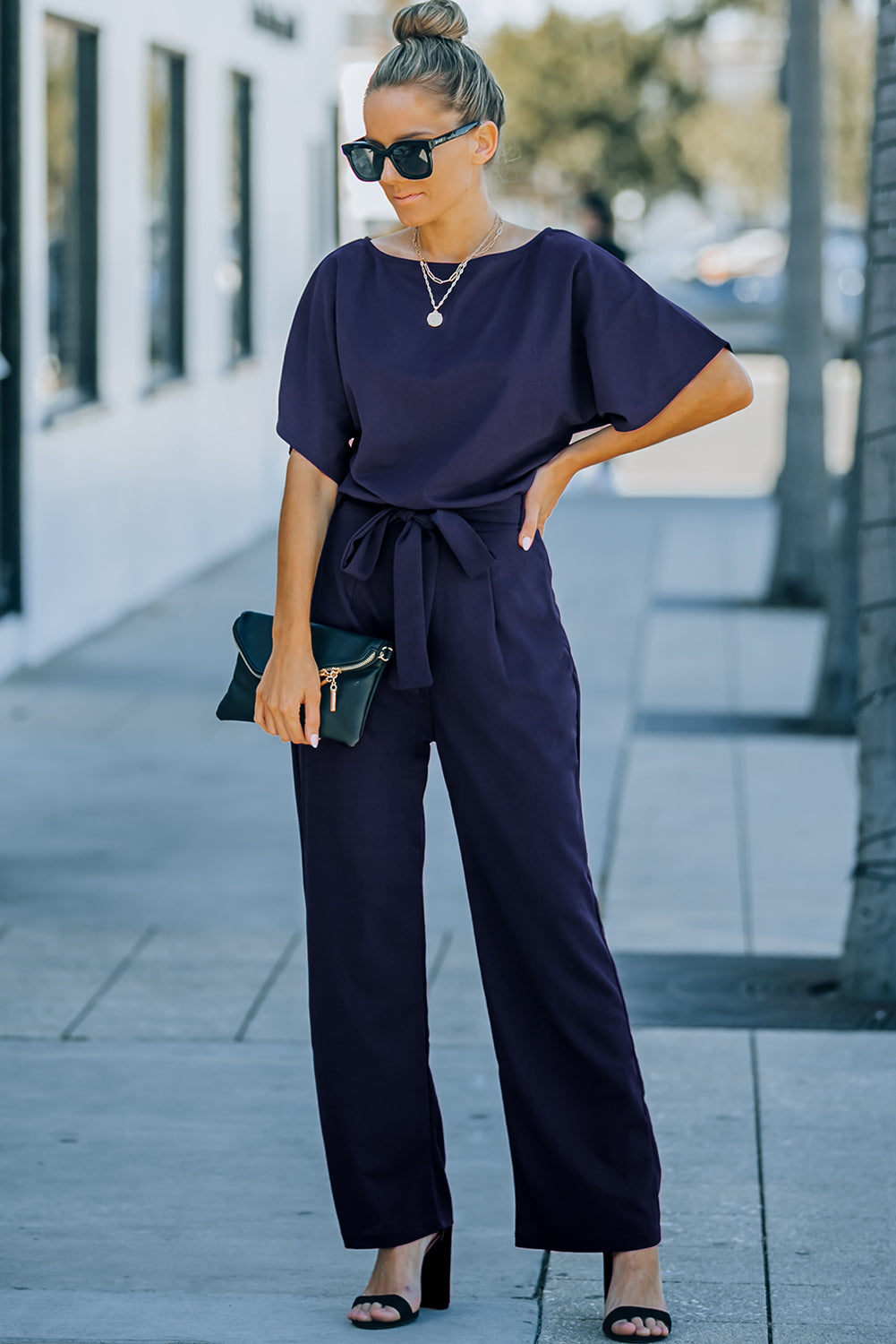 A stylish woman wearing a blue belted wide leg jumpsuit with batwing sleeves, showcasing a chic and glamorous look.