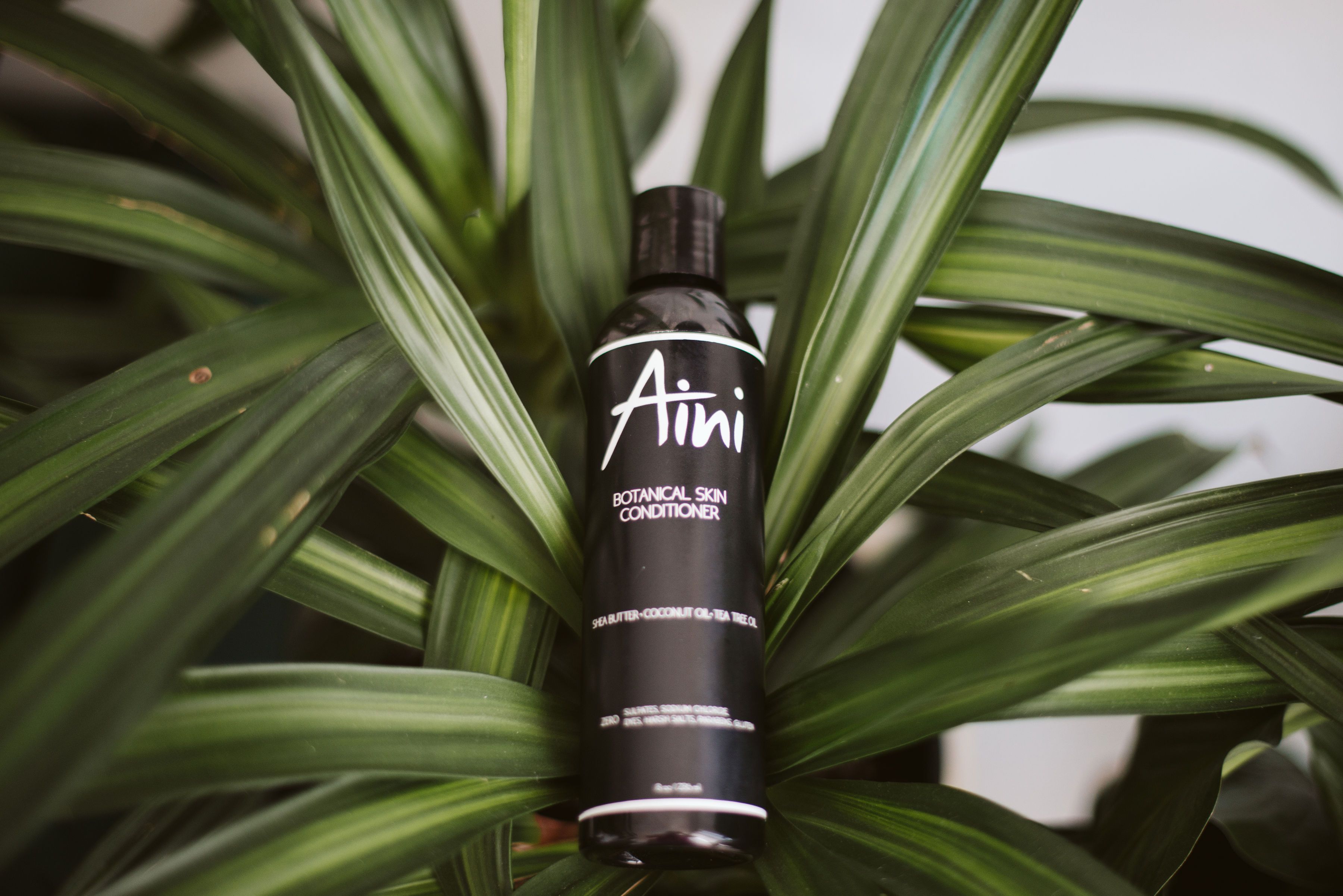 A bottle of Botanical Skin Conditioner featuring a lightweight formula with tea tree oil and shea butter, surrounded by natural ingredients like coconut oil and vanilla beans.