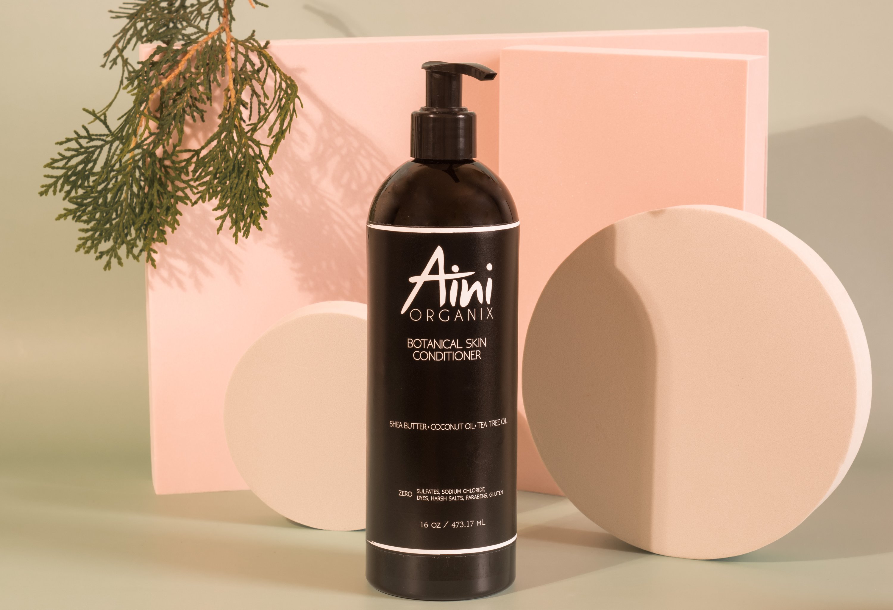 A bottle of Botanical Skin Conditioner featuring a lightweight formula with tea tree oil and shea butter, surrounded by natural ingredients like coconut oil and vanilla beans.