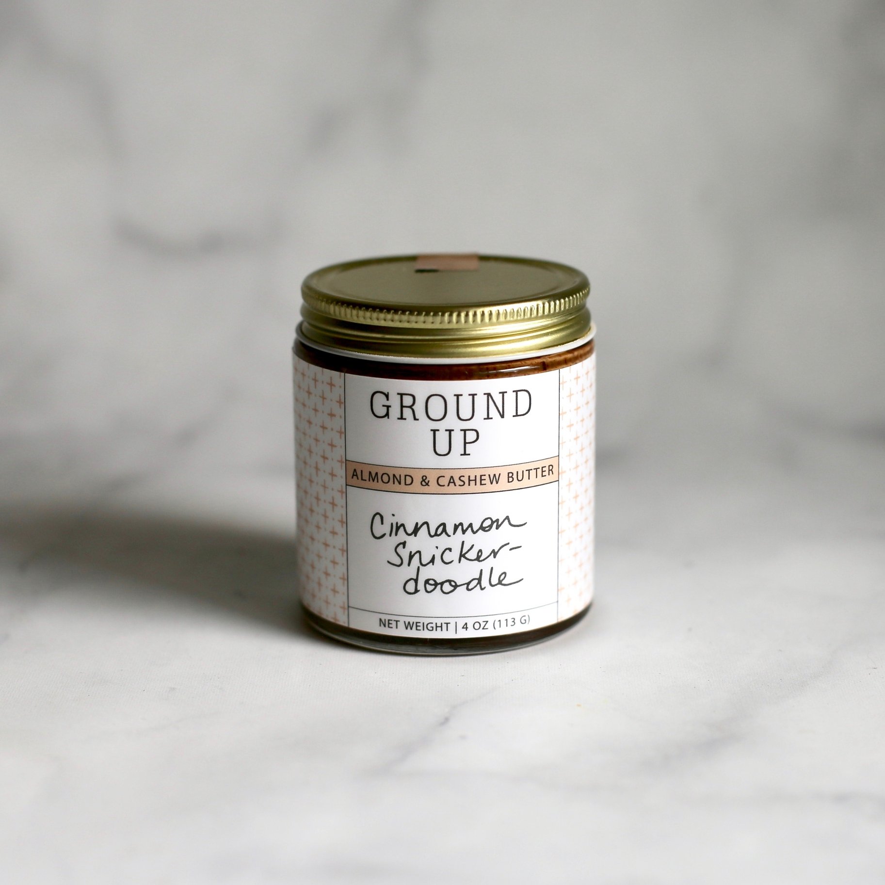 A jar of Cinnamon Snickerdoodle Almond + Cashew Nut Butter with a creamy texture and a sprinkle of cinnamon on top, showcasing its delicious flavor.