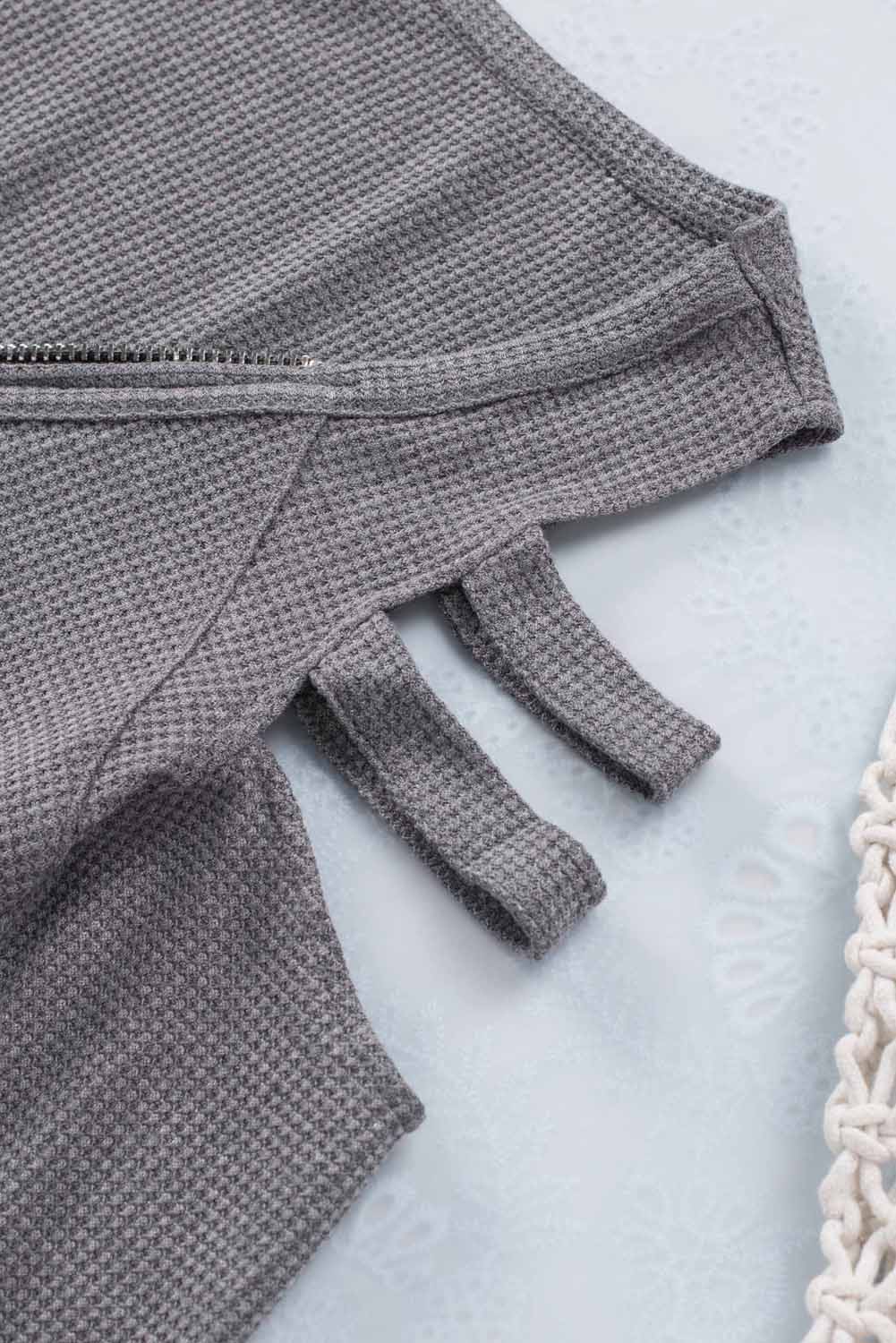 Gray long sleeve top with zip neck and cut-out design, featuring a textured waffle knit fabric and a loose fit with a ruffled hemline.