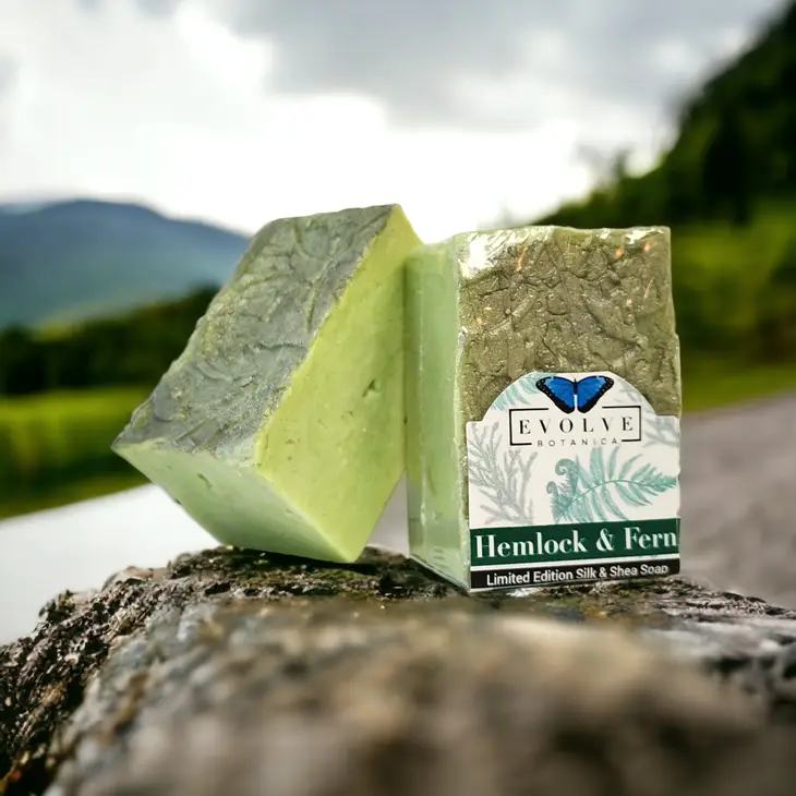 A bar of Specialty Soap - Hemlock & Fern showcasing its earthy green color and luxurious texture, surrounded by natural elements like ferns and wood.