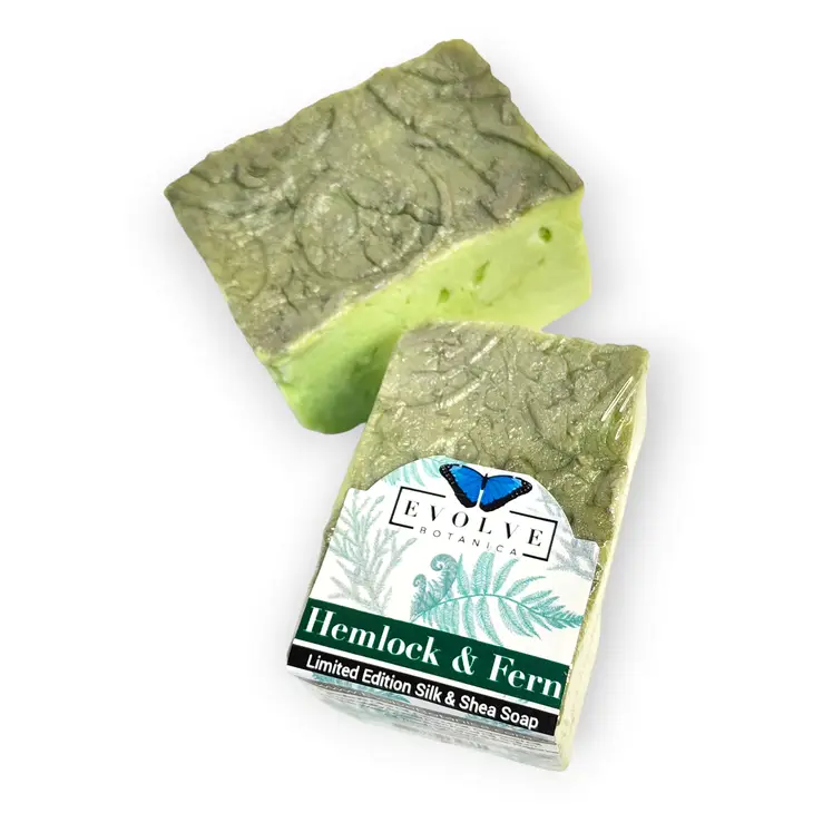 A bar of Specialty Soap - Hemlock & Fern showcasing its earthy green color and luxurious texture, surrounded by natural elements like ferns and wood.