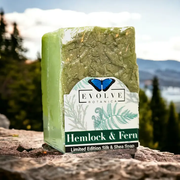 A bar of Specialty Soap - Hemlock & Fern showcasing its earthy green color and luxurious texture, surrounded by natural elements like ferns and wood.