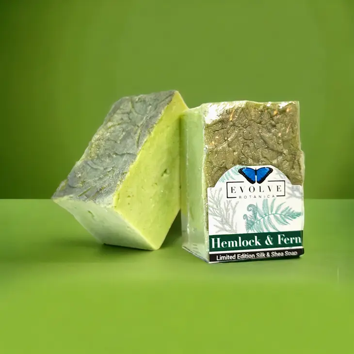 A bar of Specialty Soap - Hemlock & Fern showcasing its earthy green color and luxurious texture, surrounded by natural elements like ferns and wood.