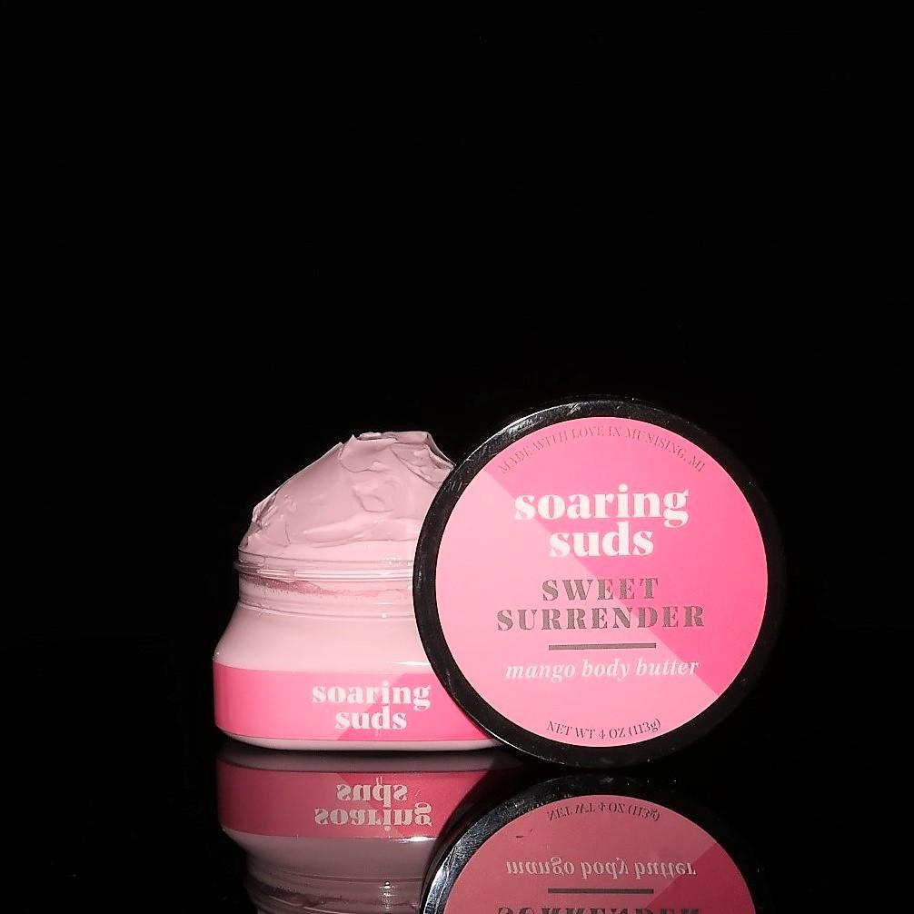 A 4 oz jar of Sweet Surrender Mango Body Butter with a creamy texture, surrounded by mangoes and strawberries, showcasing its natural ingredients.