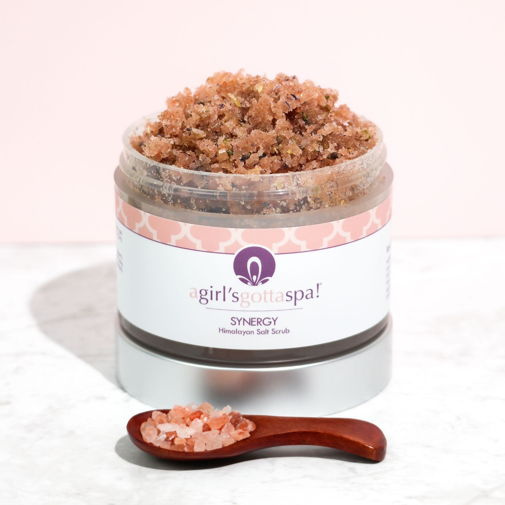 A jar of Synergy Himalayan Salt Body Scrub with a wooden spoon, showcasing its natural pink salt texture and aromatic ingredients.