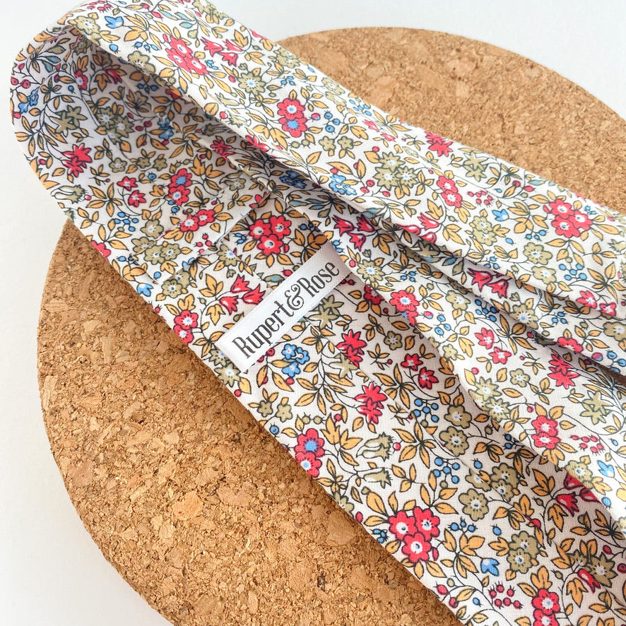 RUBEN Boys Self Tie featuring a floral pattern in sage green, cornflower blue, yellow, and vibrant pink on an off-white background.