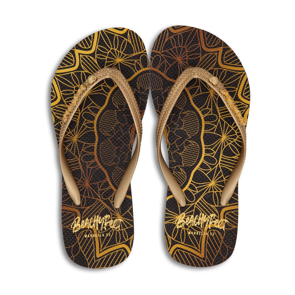 BeachyFeet® Mandala flip flops featuring a black and gold bohemian print with an orange strap and golden emblem.