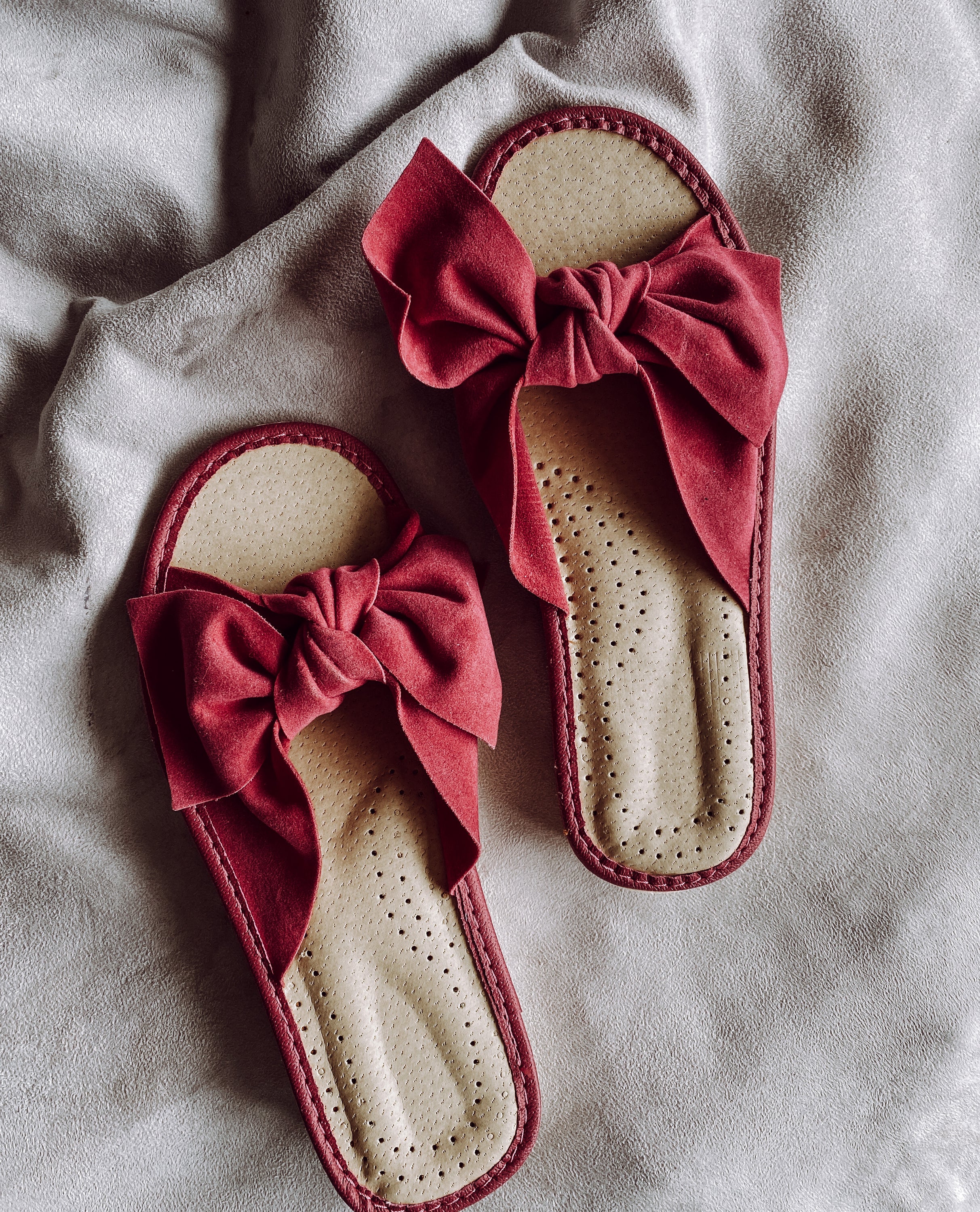 A pair of pink suede slippers featuring a decorative ribbon on the front, designed for comfort with a cushioned insole and foam bottom.