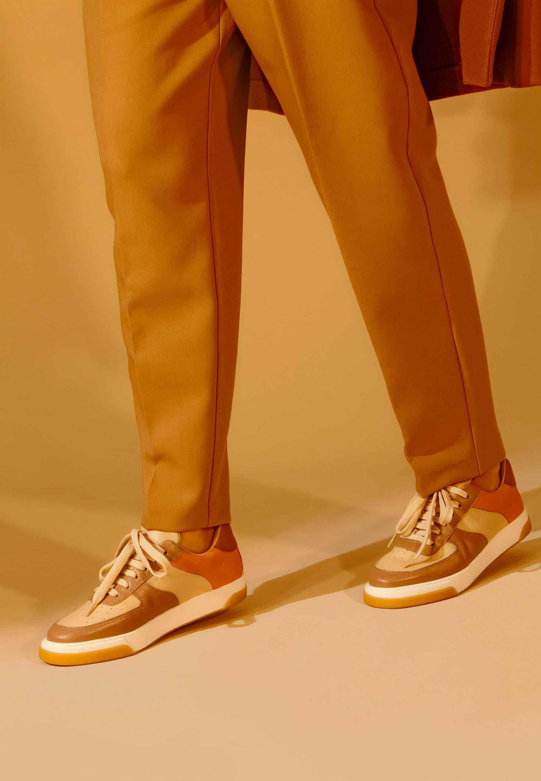 A pair of elegant women's sneakers in beige leather with brown and orange accents, featuring a lace-up front and padded collar.