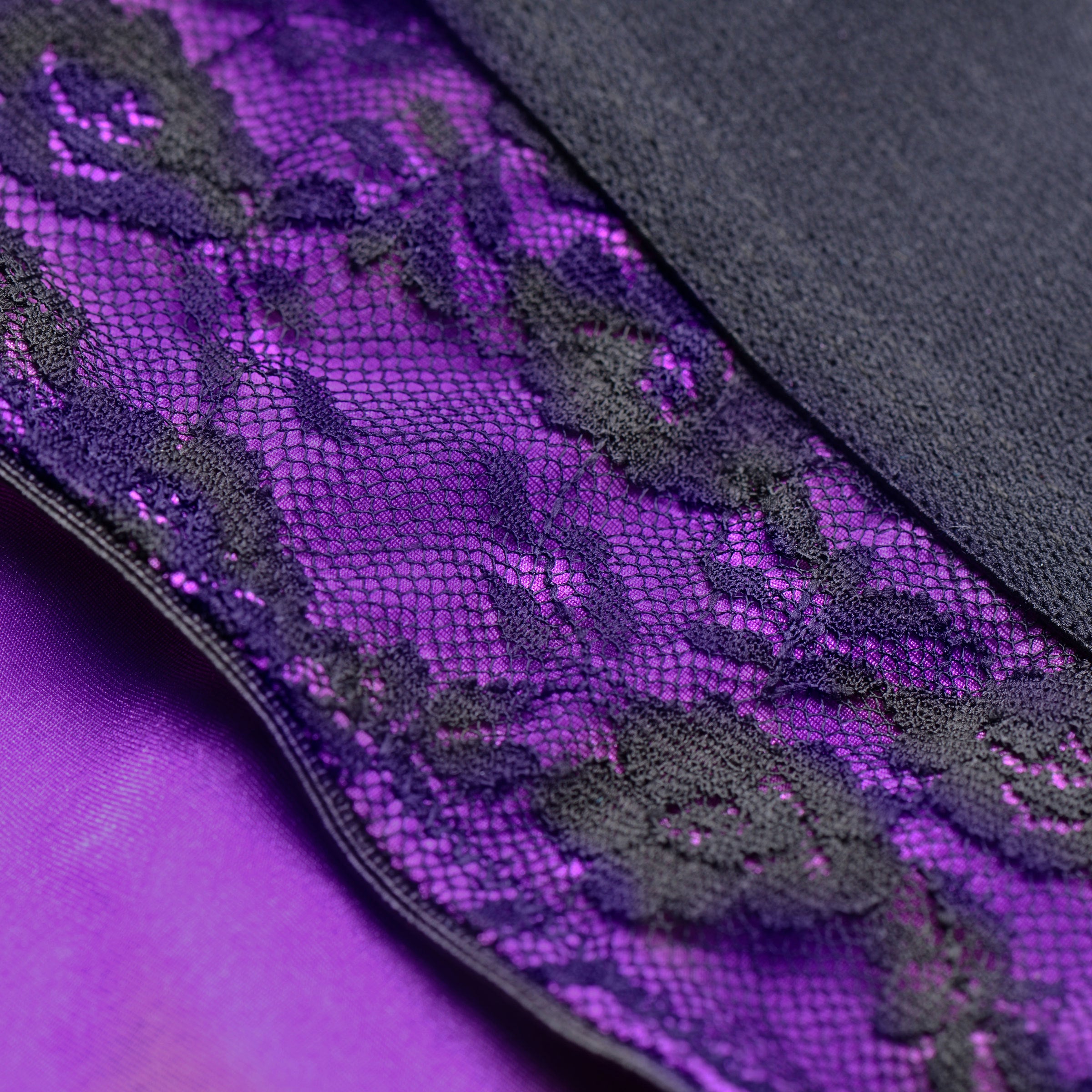 Lace Envy Crotchless Panty Harness in purple and black, featuring a wide elastic strap and universal O-ring for dildos.