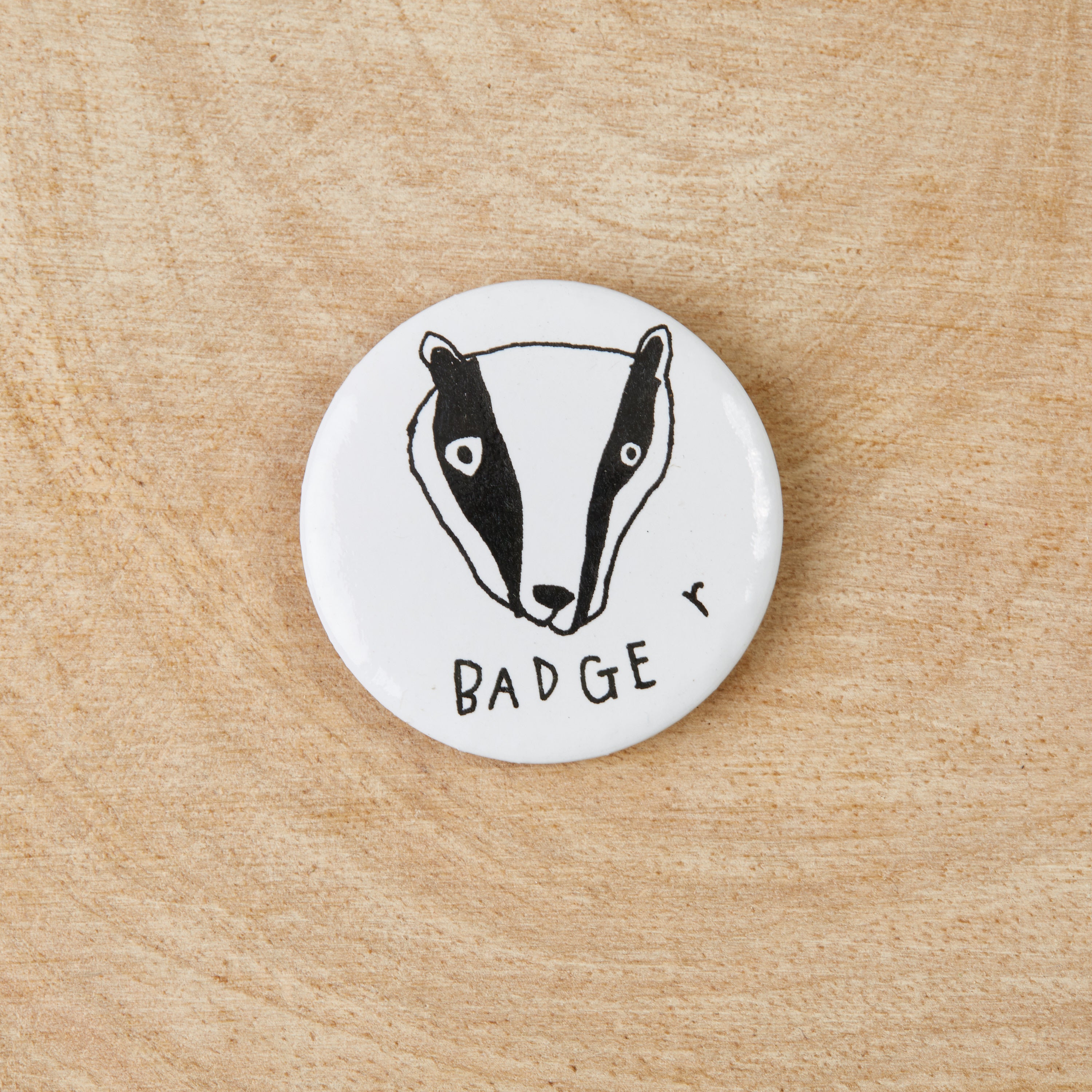 A white button badge featuring a black badger design, measuring 38mm in width, made from 100% recycled materials.