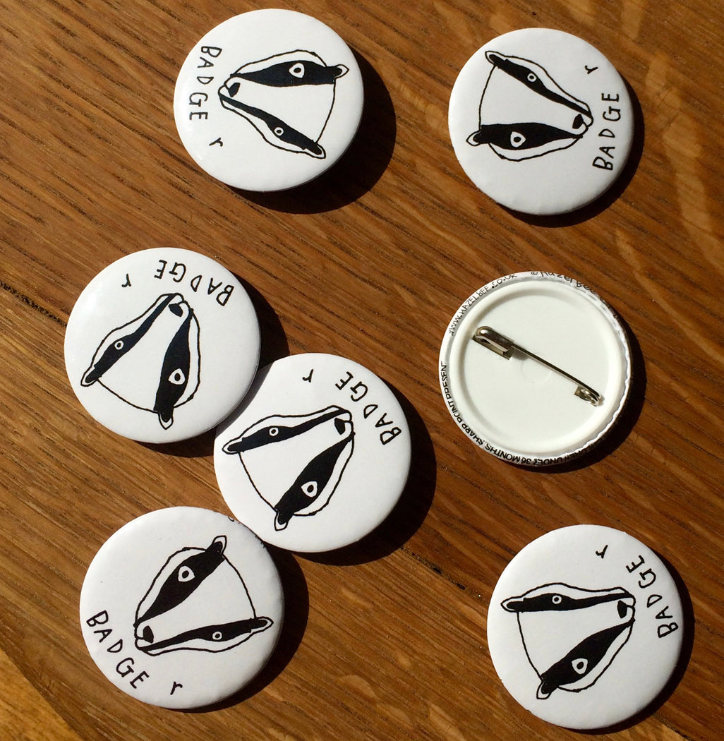 A white button badge featuring a black badger design, measuring 38mm in width, made from 100% recycled materials.