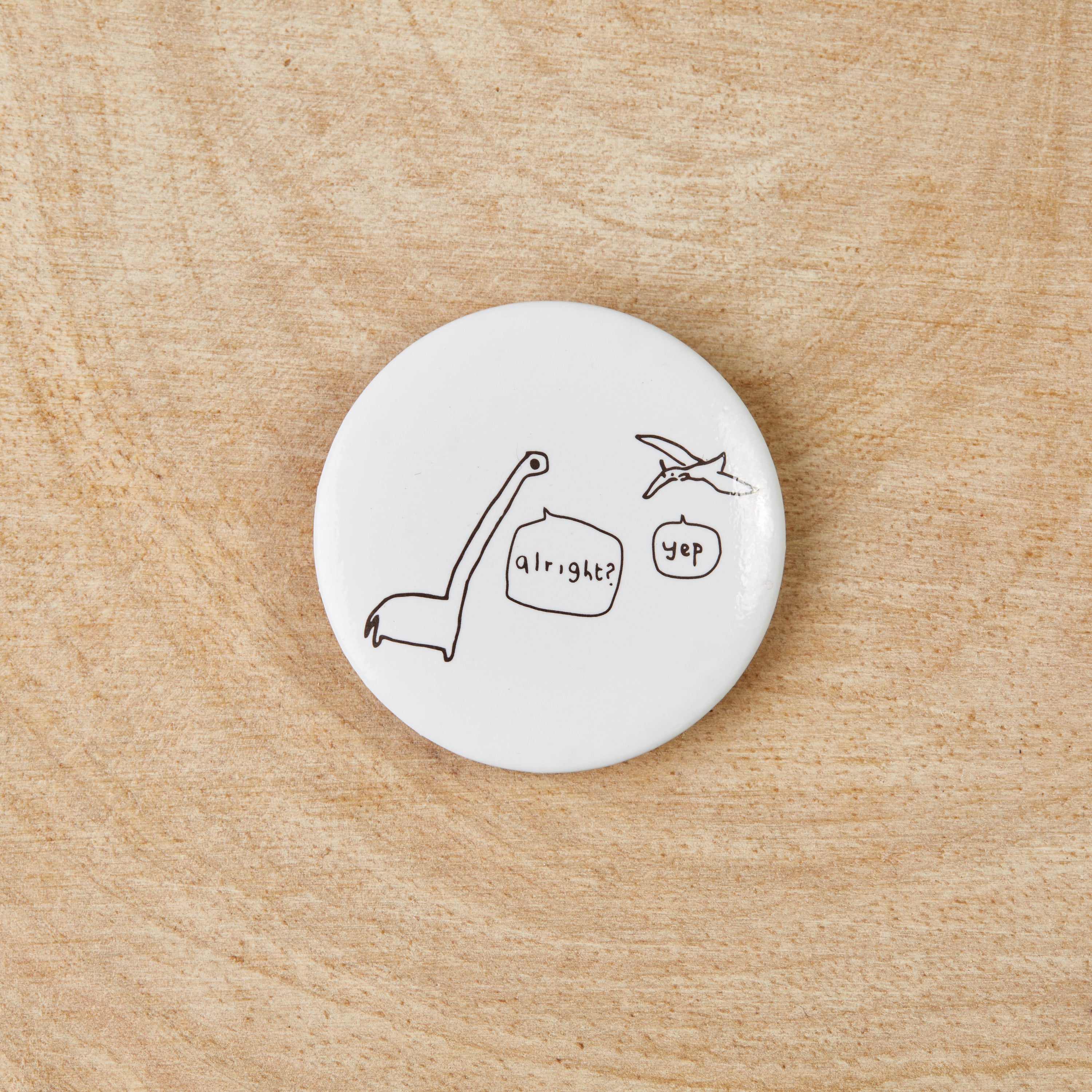 A white button badge featuring a black dinosaur design, measuring 38mm in width, made from recycled materials.