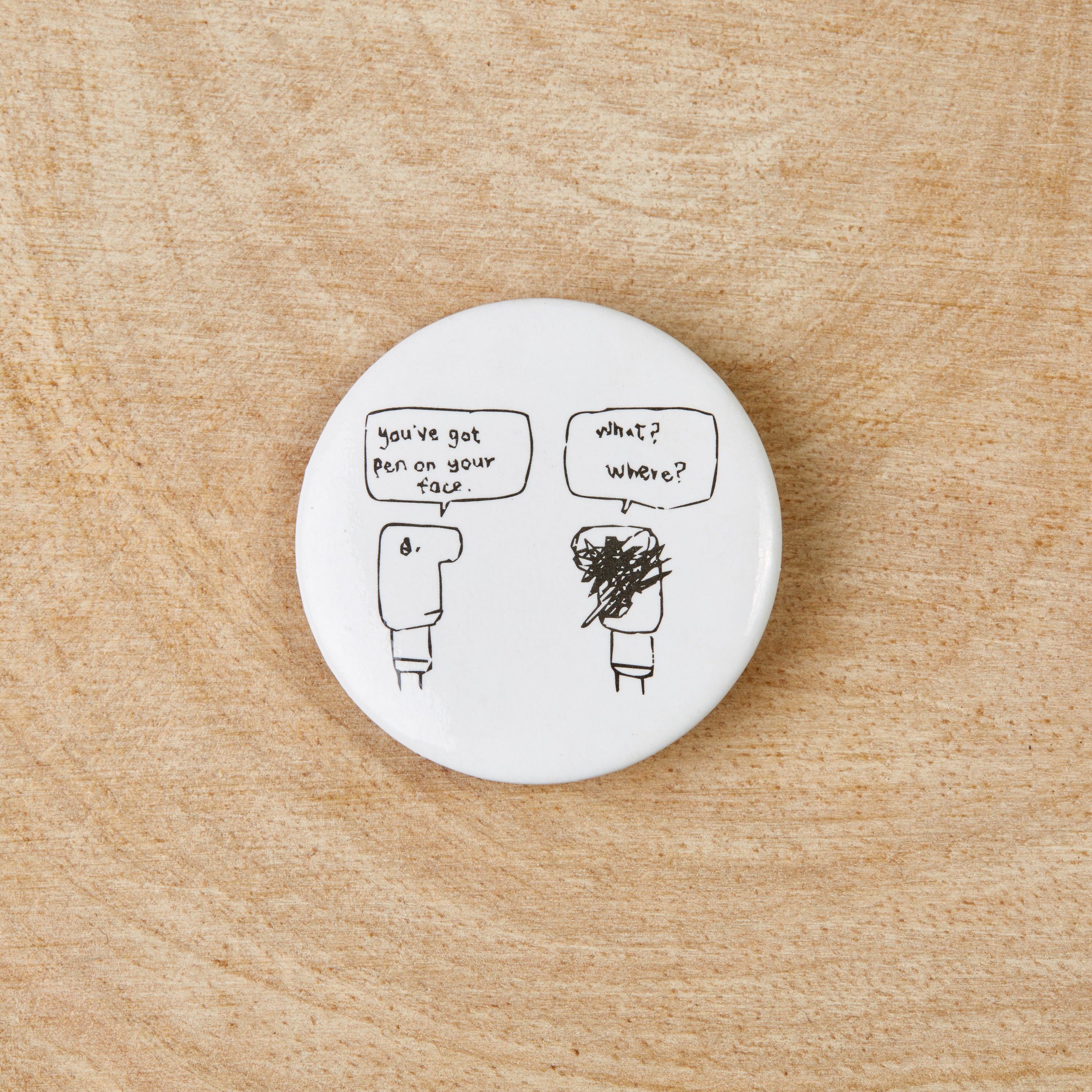 A white button badge featuring a black design of a pen on a face, measuring 38mm in diameter, made from recycled materials.