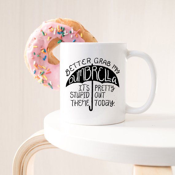 Funny coffee mug with the phrase 'Better Grab My Dumbrella... It's Pretty Stupid Out' printed on it, showcasing a humorous design.