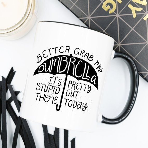 Funny coffee mug with the phrase 'Better Grab My Dumbrella... It's Pretty Stupid Out' printed on it, showcasing a humorous design.