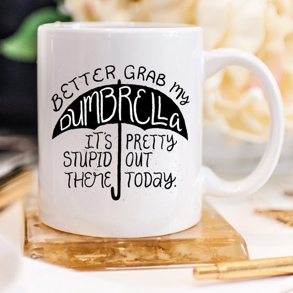 Funny coffee mug with the phrase 'Better Grab My Dumbrella... It's Pretty Stupid Out' printed on it, showcasing a humorous design.