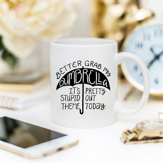 Funny coffee mug with the phrase 'Better Grab My Dumbrella... It's Pretty Stupid Out' printed on it, showcasing a humorous design.