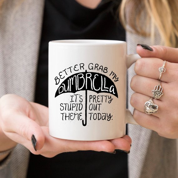 Funny coffee mug with the phrase 'Better Grab My Dumbrella... It's Pretty Stupid Out' printed on it, showcasing a humorous design.