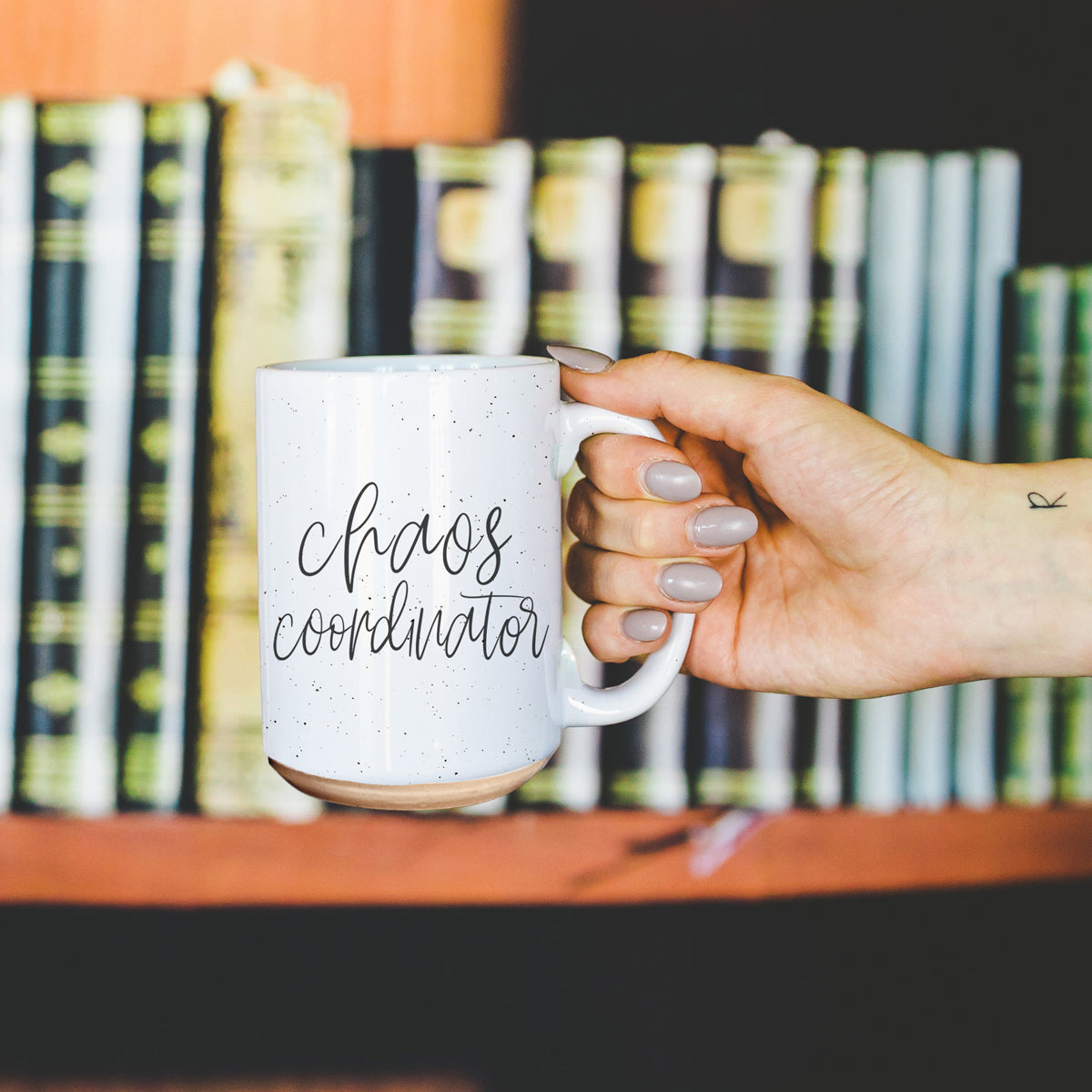 A humorous 'Chaos Coordinator' 16oz coffee mug featuring a black graphic on an off-white ceramic body with dark speckles.